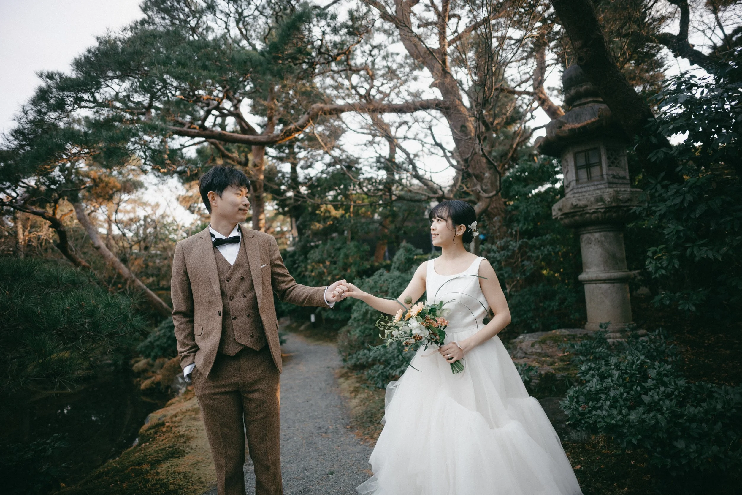 A bride and groom holding hands in a garden with trees and a stone lantern.