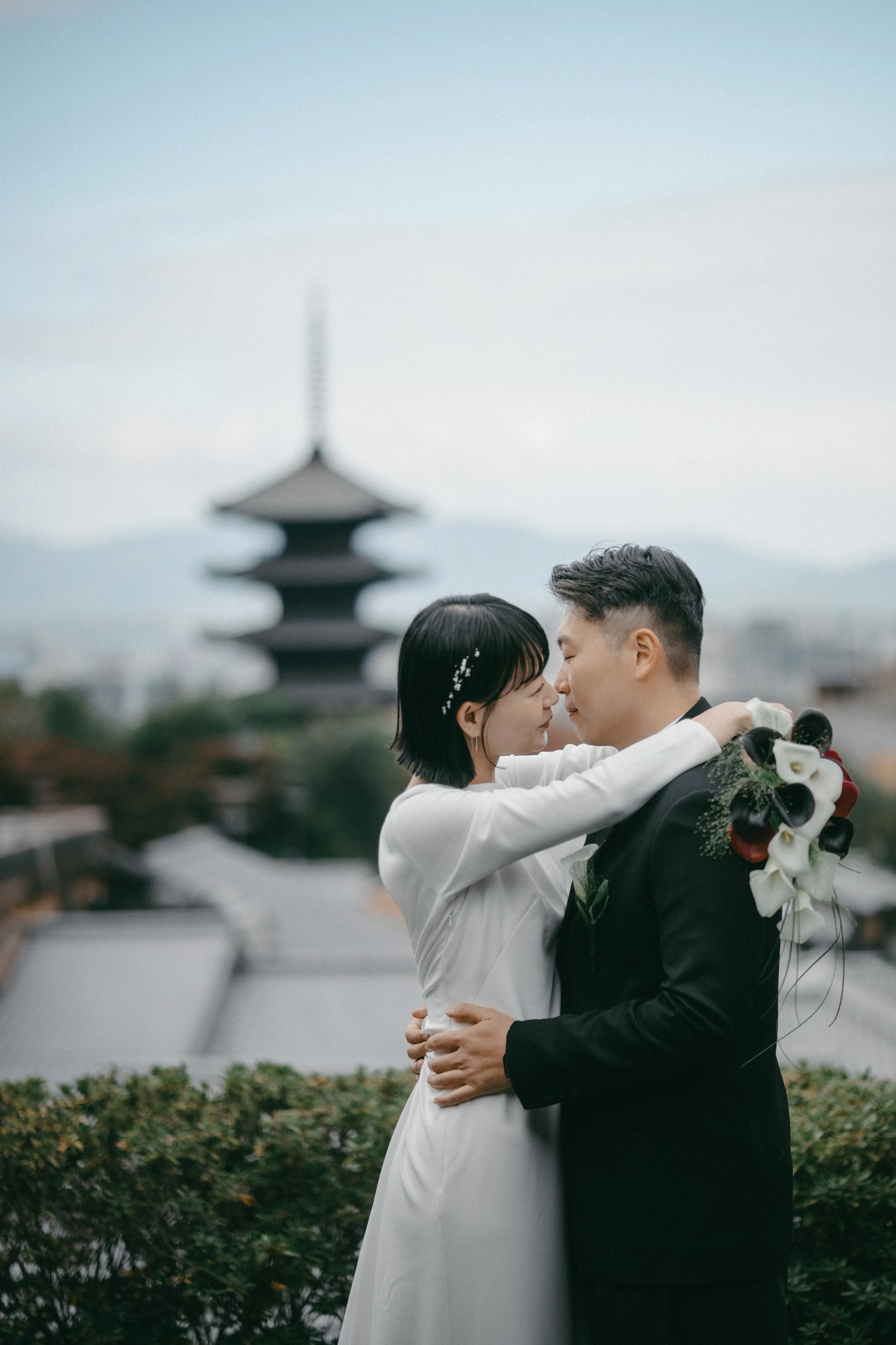 A bride and groom embrace on a rooftop with a pagoda in the background, the bride holding a bouquet, and they are closing their eyes.