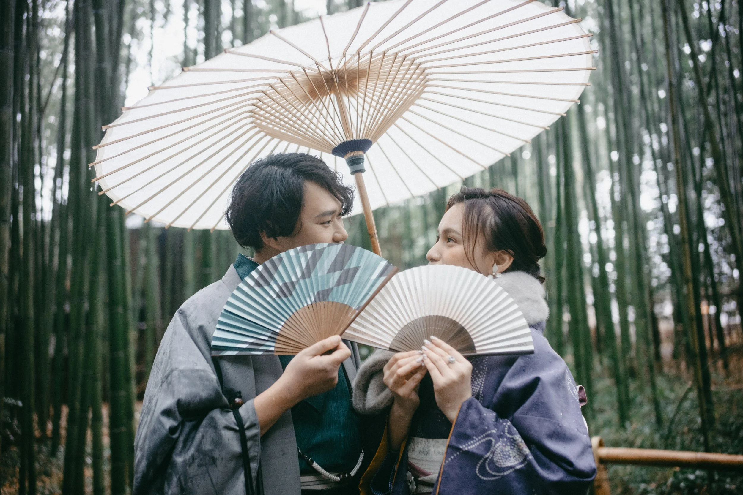 Two women in traditional Japanese kimonos holding paper fans and standing close together in a bamboo forest, with two umbrellas overhead.