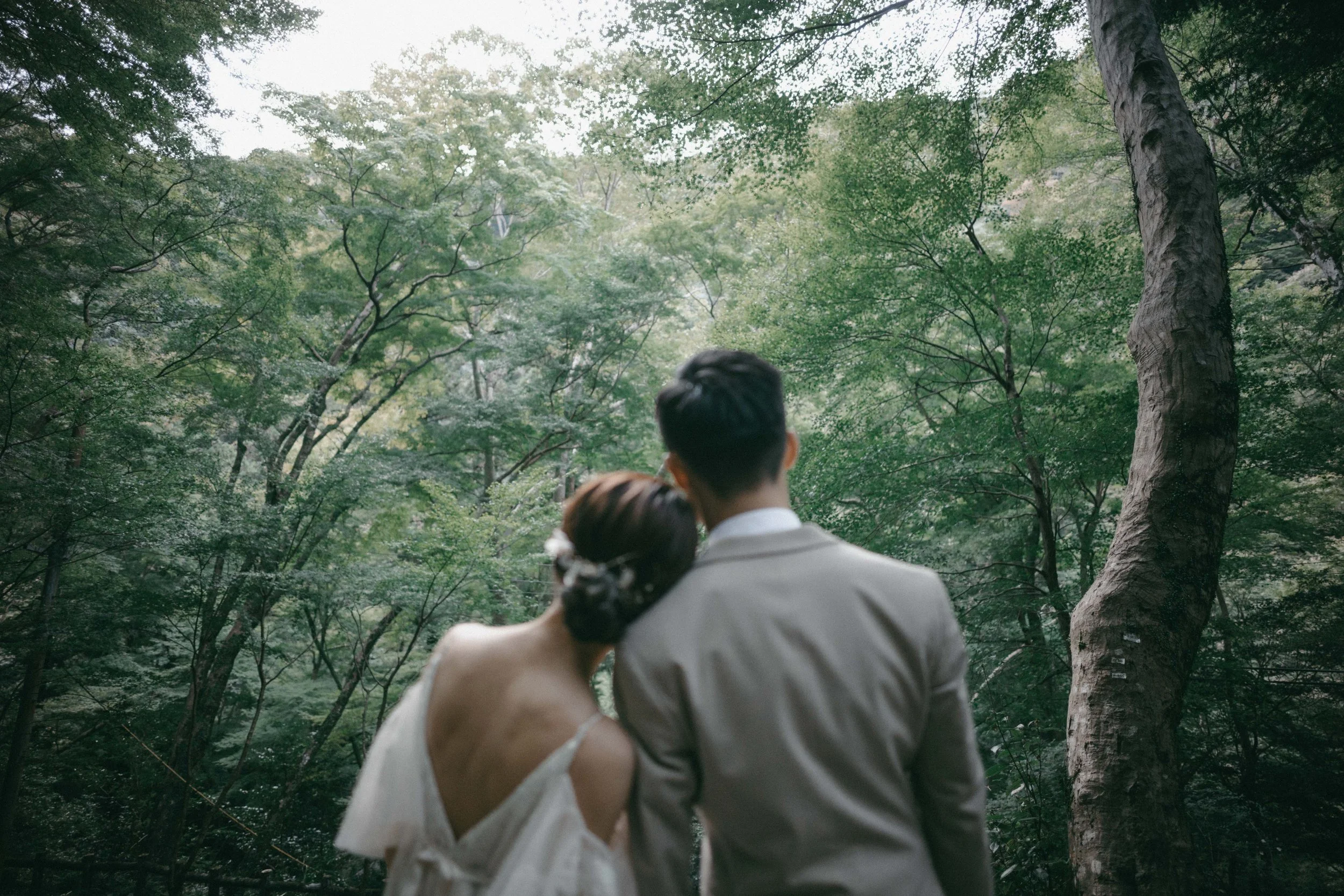 A couple, dressed in wedding attire, standing close together in a lush green forest, with their backs to the camera.