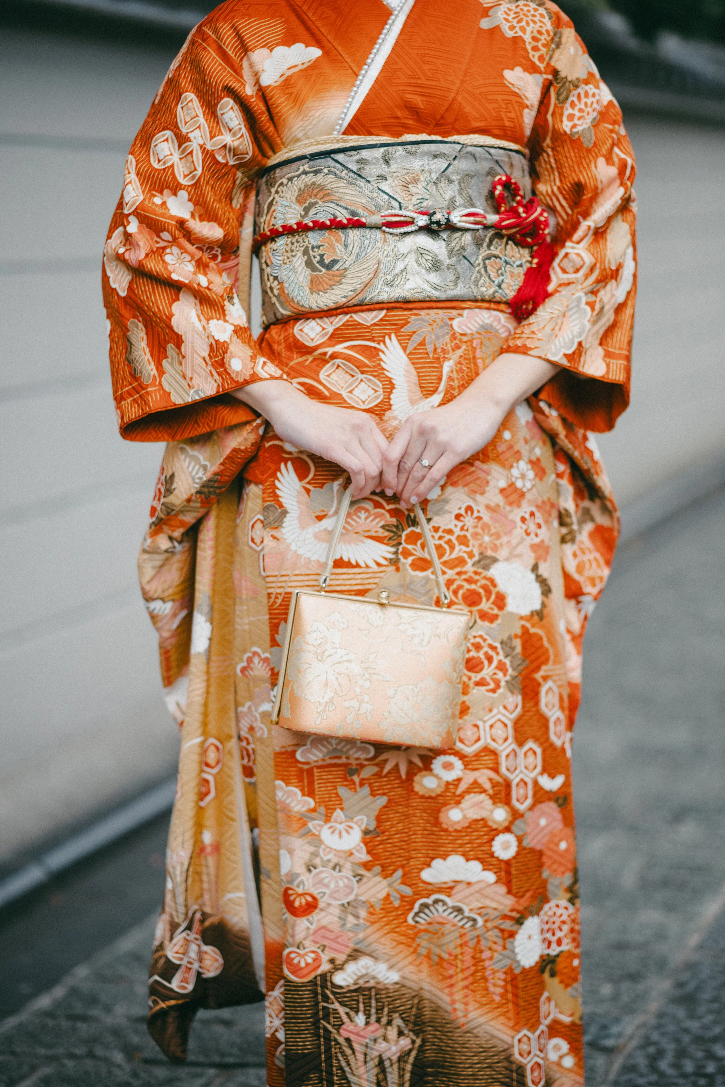 A person wearing a traditional orange kimono with floral and crane patterns, holding a gold clutch, standing on a street.