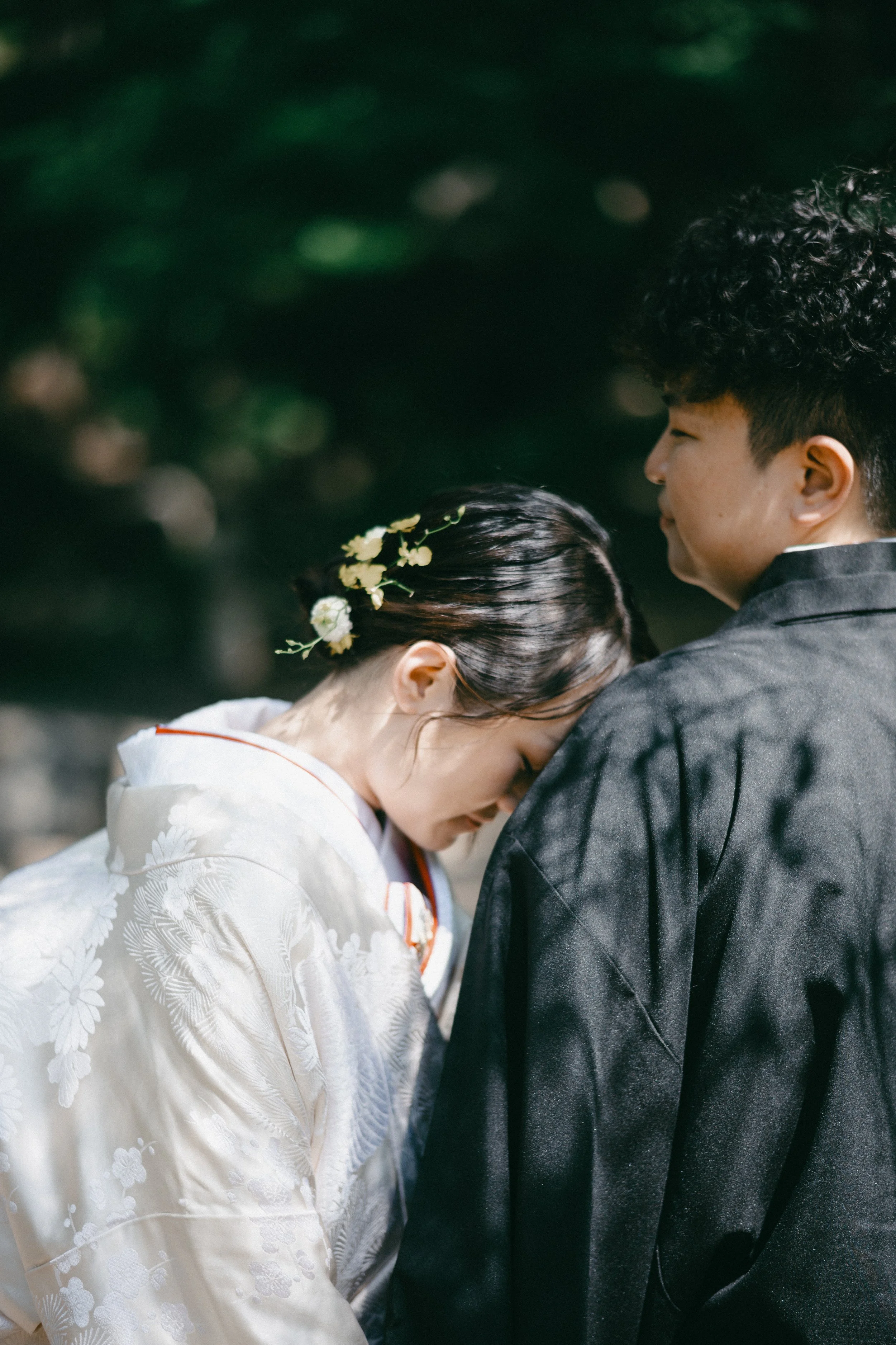 A woman in traditional Japanese wedding attire with flowers in her hair leaning her forehead against a man in a dark kimono outdoors.