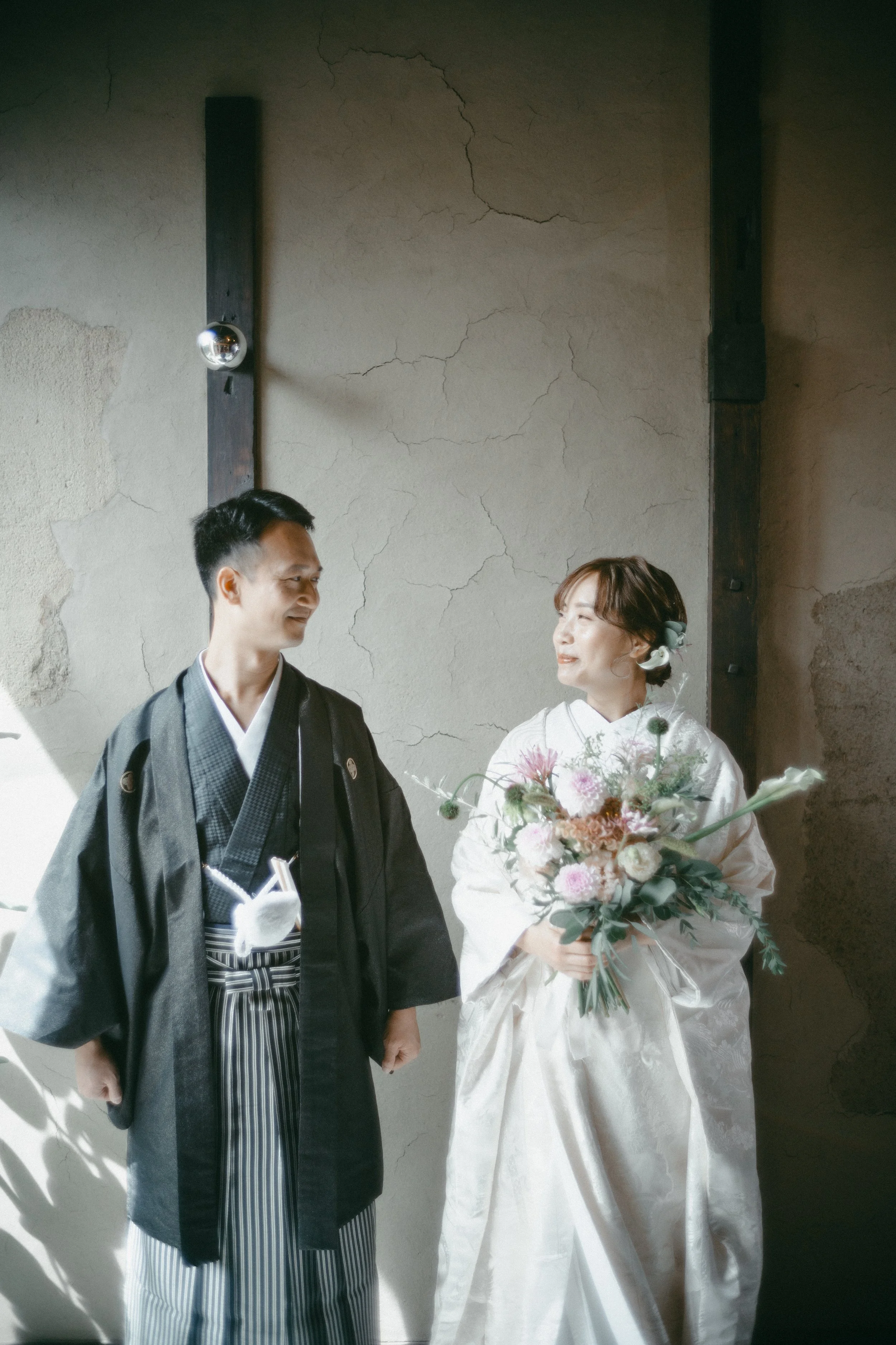A bride in a white wedding kimono holding a bouquet, standing and smiling at a groom in traditional black kimono with striped hakama, indoors beside a textured wall.