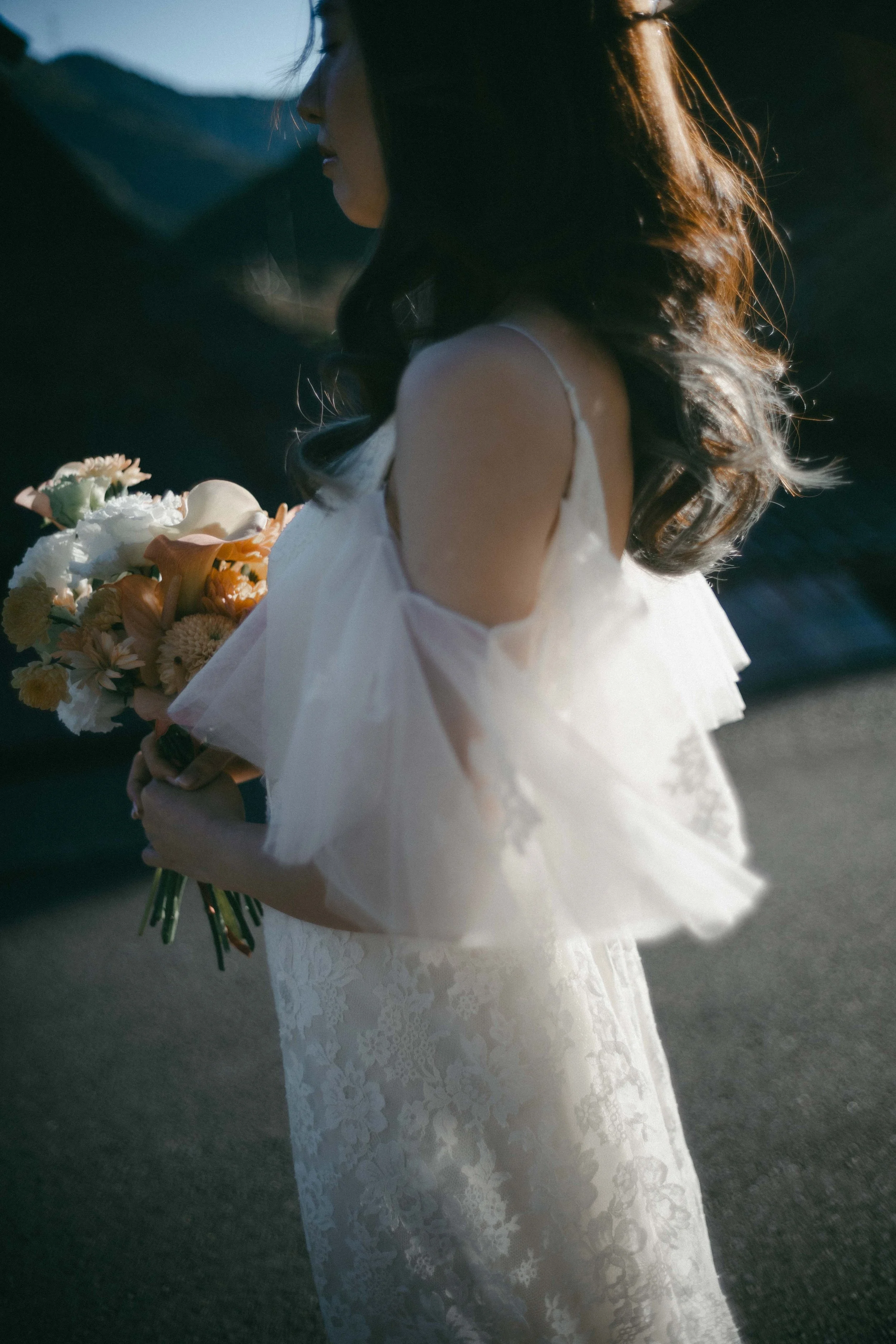 A woman in a white lace dress holding a bouquet of flowers on a dark outdoor setting.