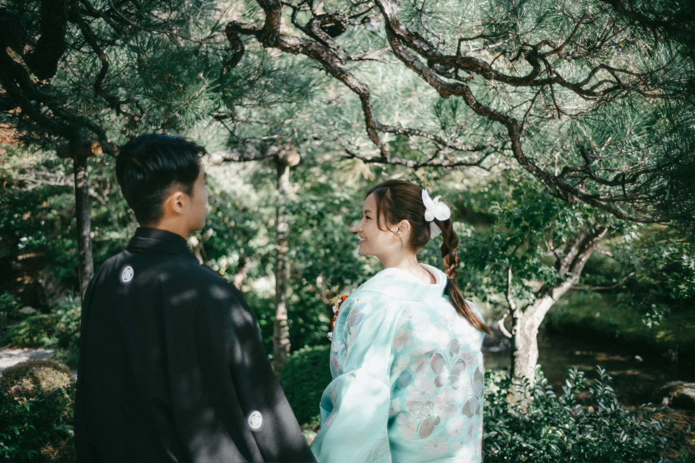 A young couple dressed in traditional Japanese attire enjoying a moment together in a lush, green garden under a tree.