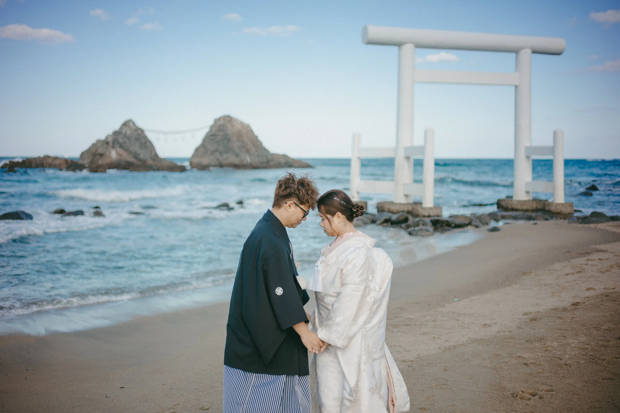 A couple dressed in traditional Japanese wedding attire holding hands on a beach with a torii gate in the background.