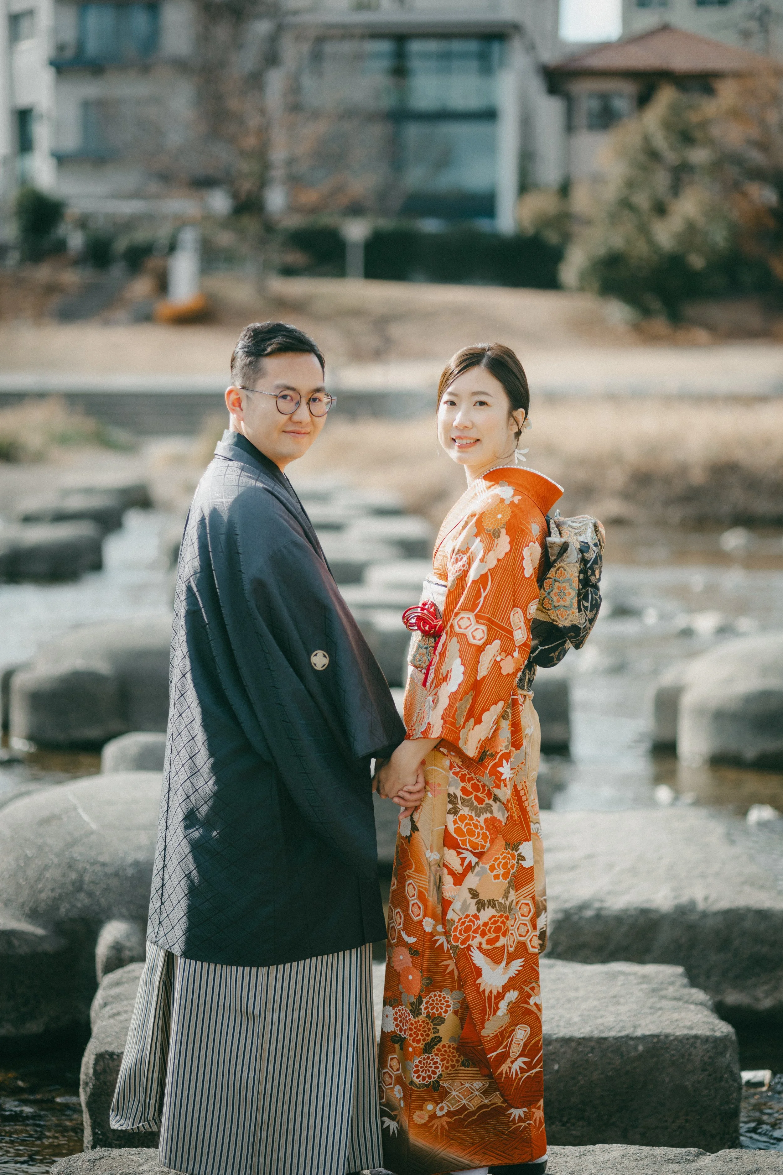 A couple dressed in traditional Japanese kimonos holding hands outdoors near a river with rocks, with trees and buildings in the background.
