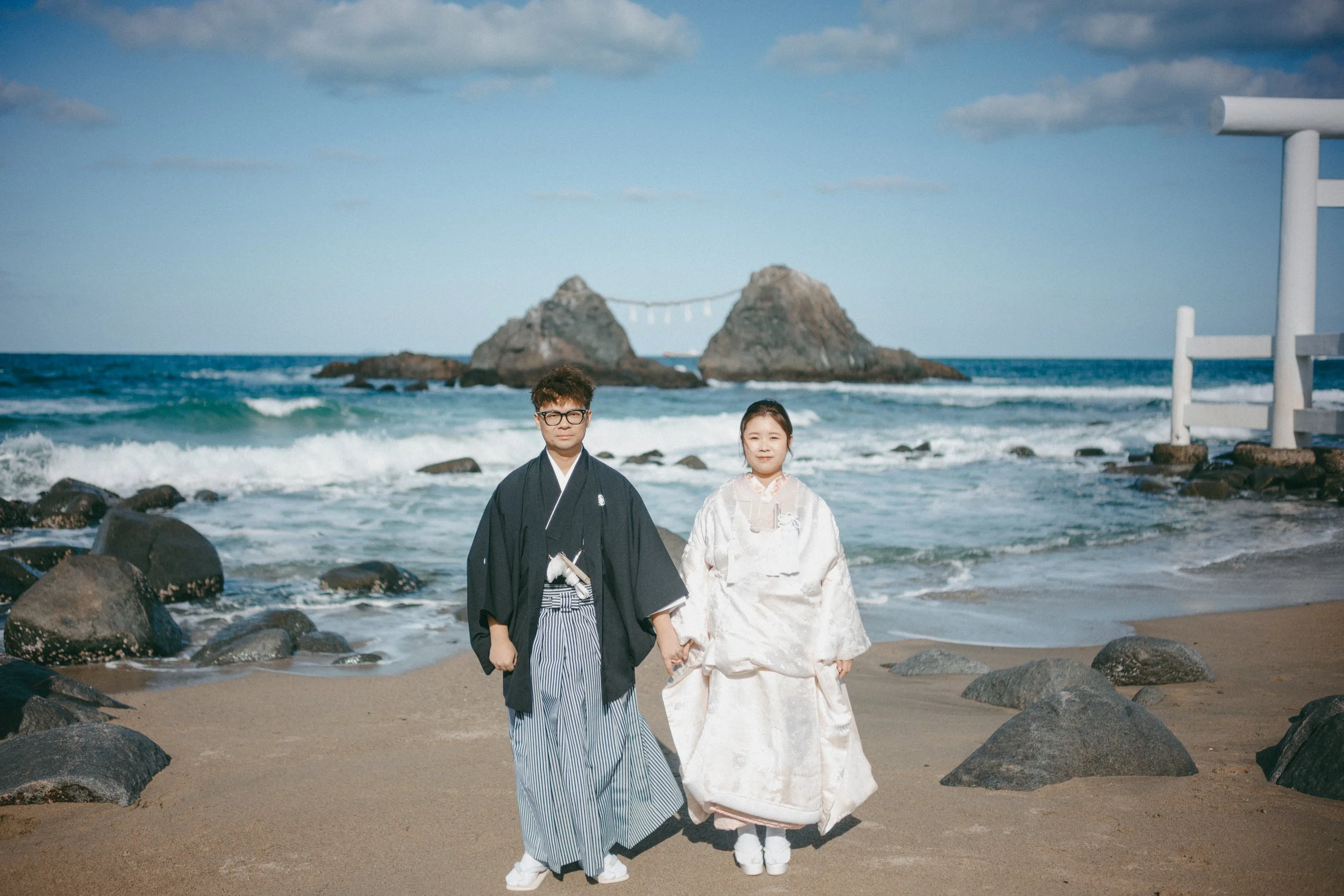 A couple dressed in traditional Japanese wedding attire walking hand-in-hand on a beach with rocks, ocean waves, and two large rocks formation with a shinto shrine in the distance.