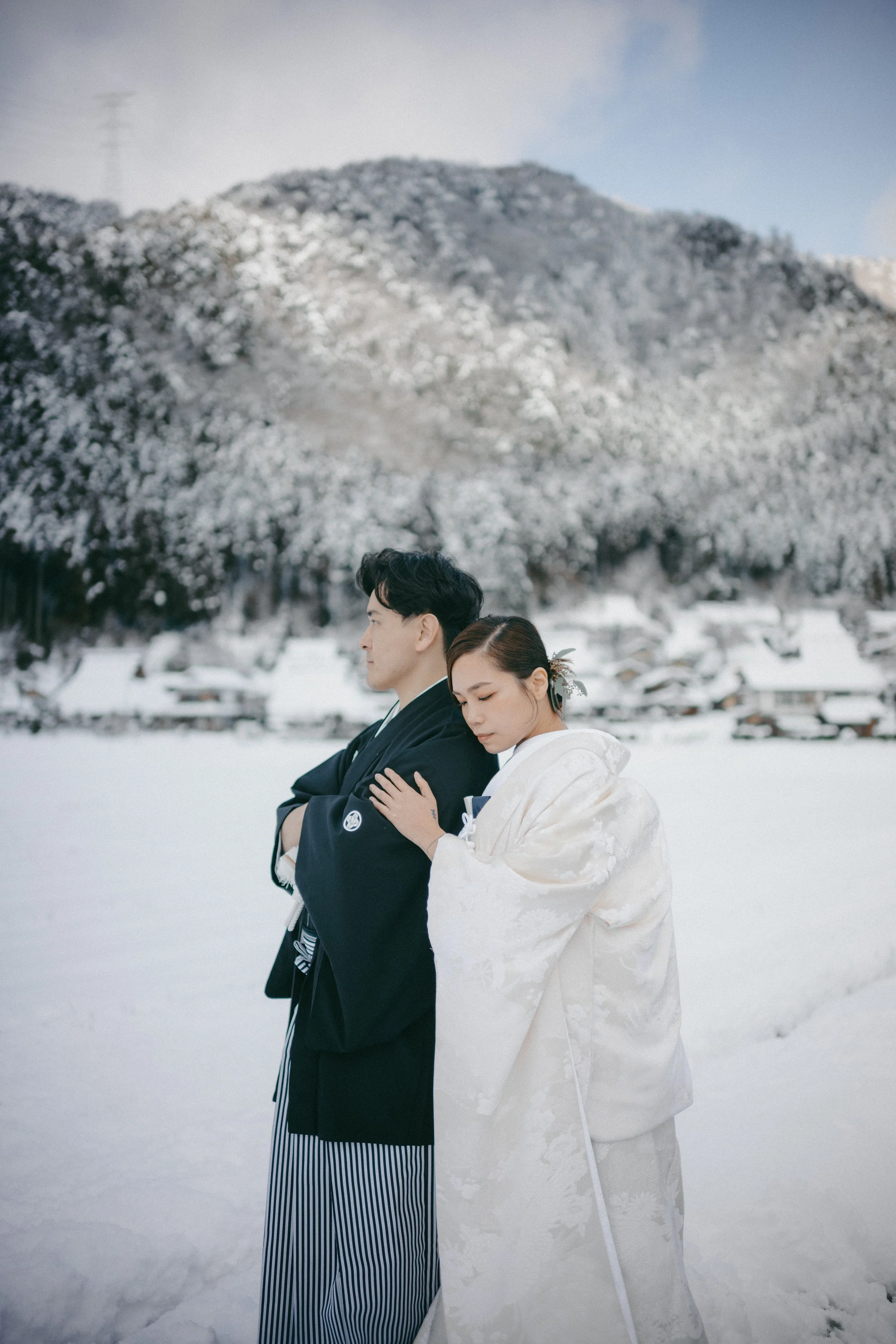 A man and woman in traditional Japanese wedding attire standing close in a snowy landscape with snow-covered houses and a mountain in the background.