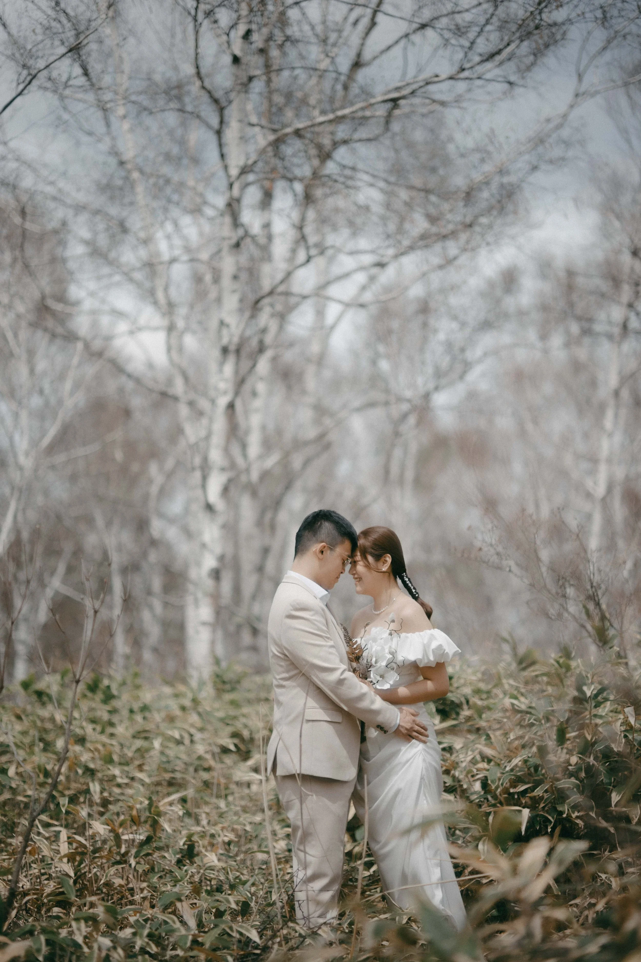 A couple in wedding attire standing close together in a forest with bare trees, holding each other with foreheads touching, in a tender moment.