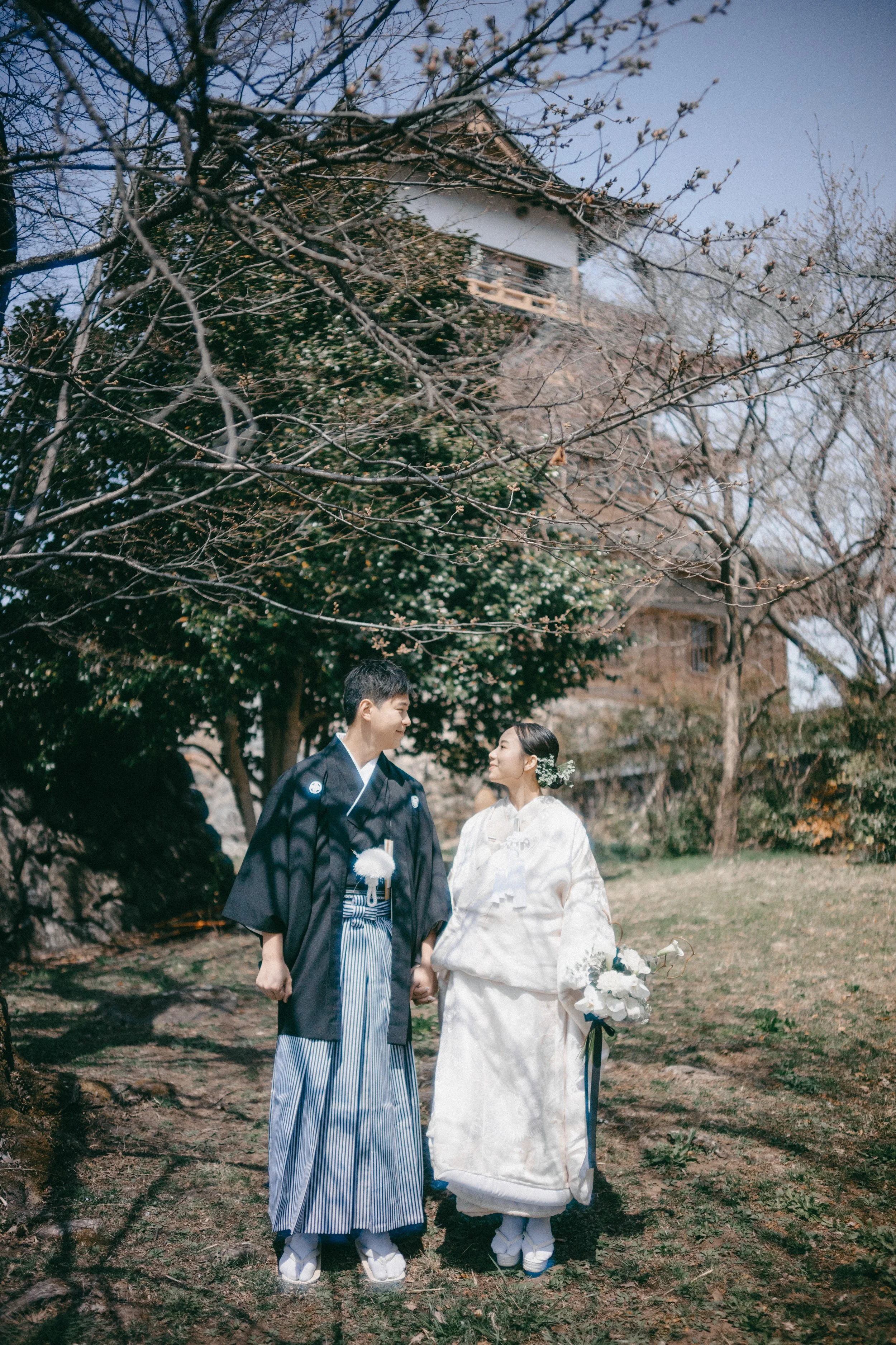 A Japanese bride and groom in traditional wedding attire standing outdoors beneath trees with a historic building in the background.
