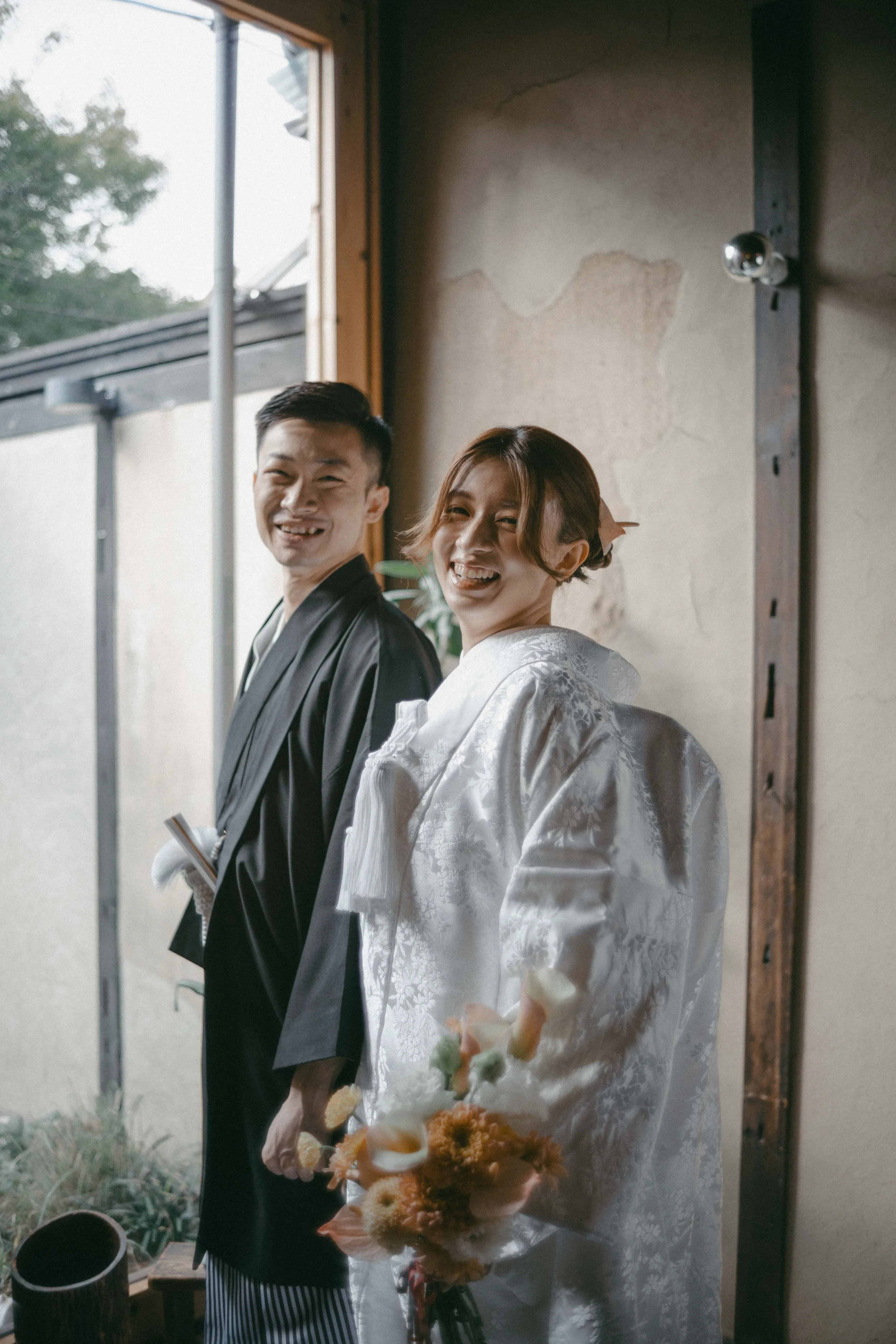 A couple in traditional Japanese wedding attire smiling indoors, with the woman holding a bouquet of flowers.