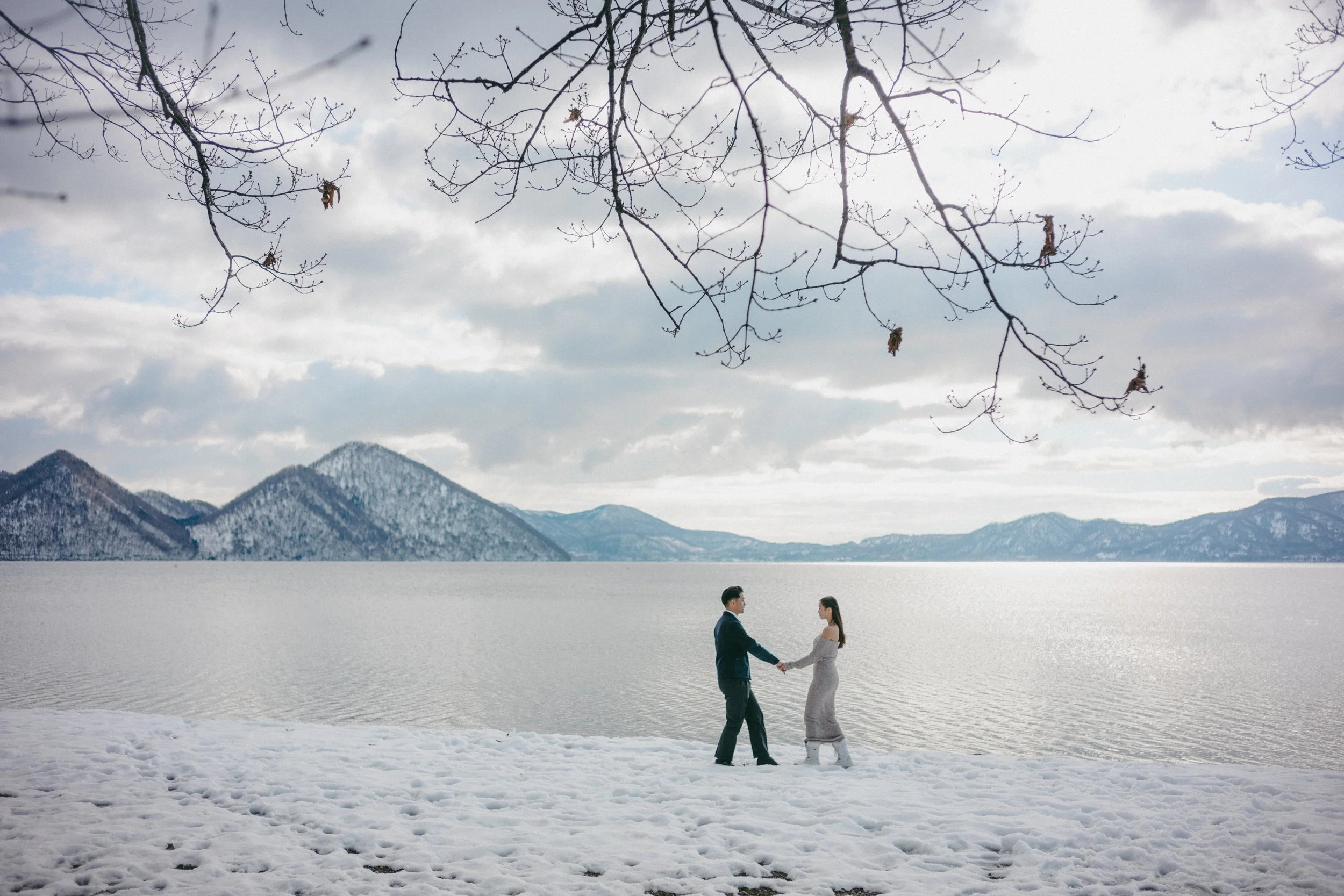 A couple standing hand in hand on snow-covered ground beside a large body of water with mountains in the background on a cloudy day.