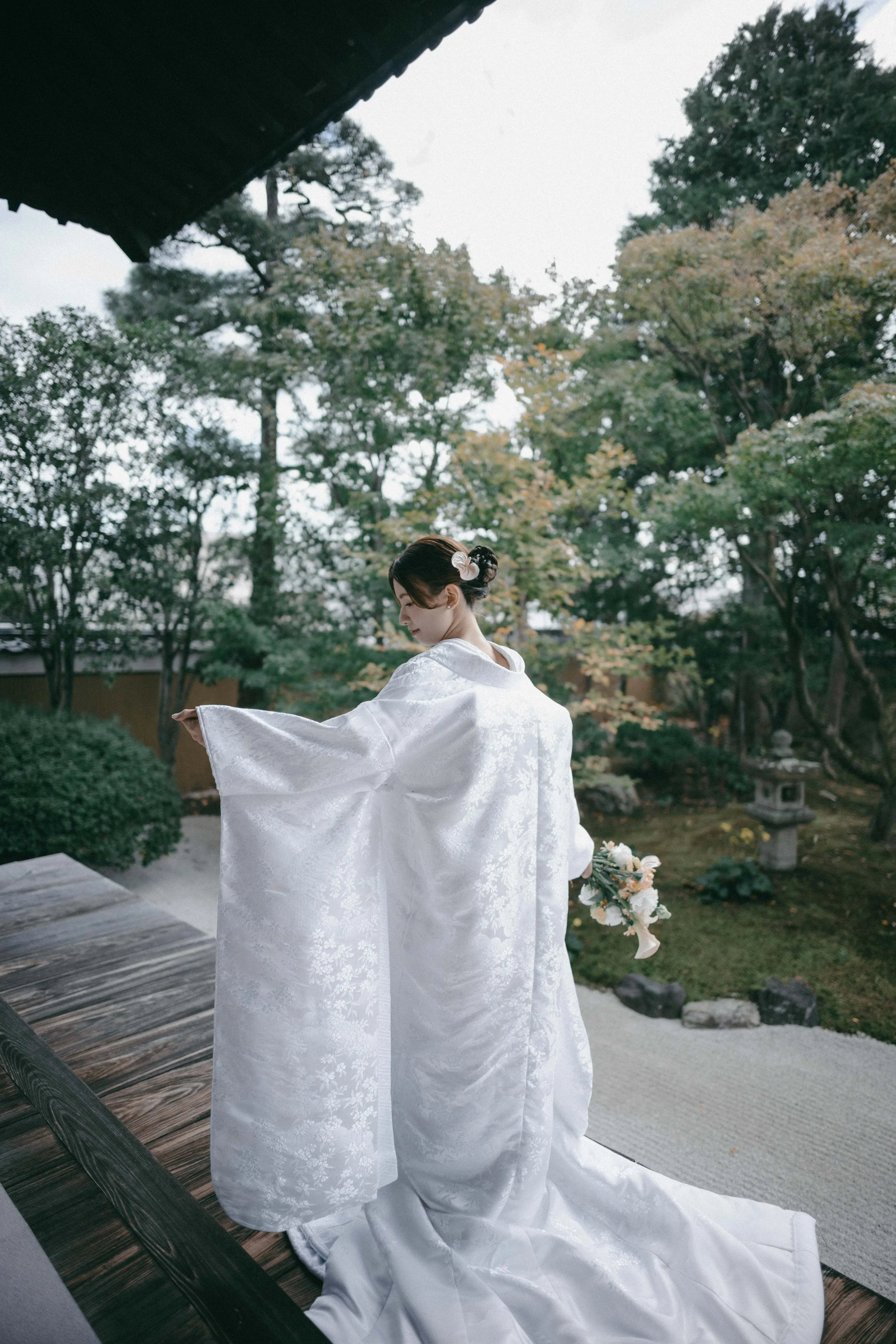 A woman in a traditional white wedding kimono, holding a bouquet, standing on a wooden porch in a Japanese garden with trees and stone lanterns.
