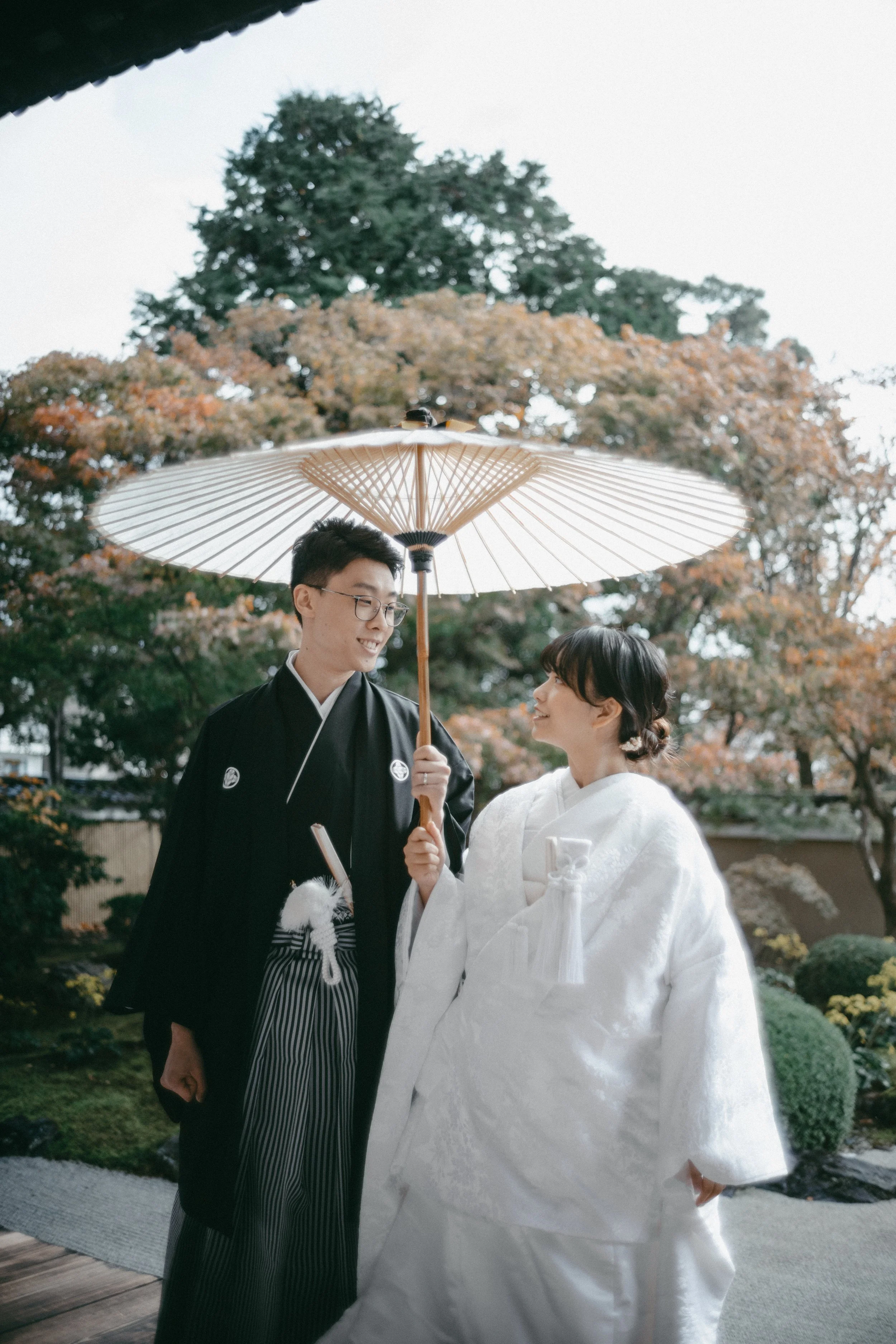 A bride and groom in traditional Japanese wedding attire standing outside under a parasol, surrounded by autumn trees and garden scenery.