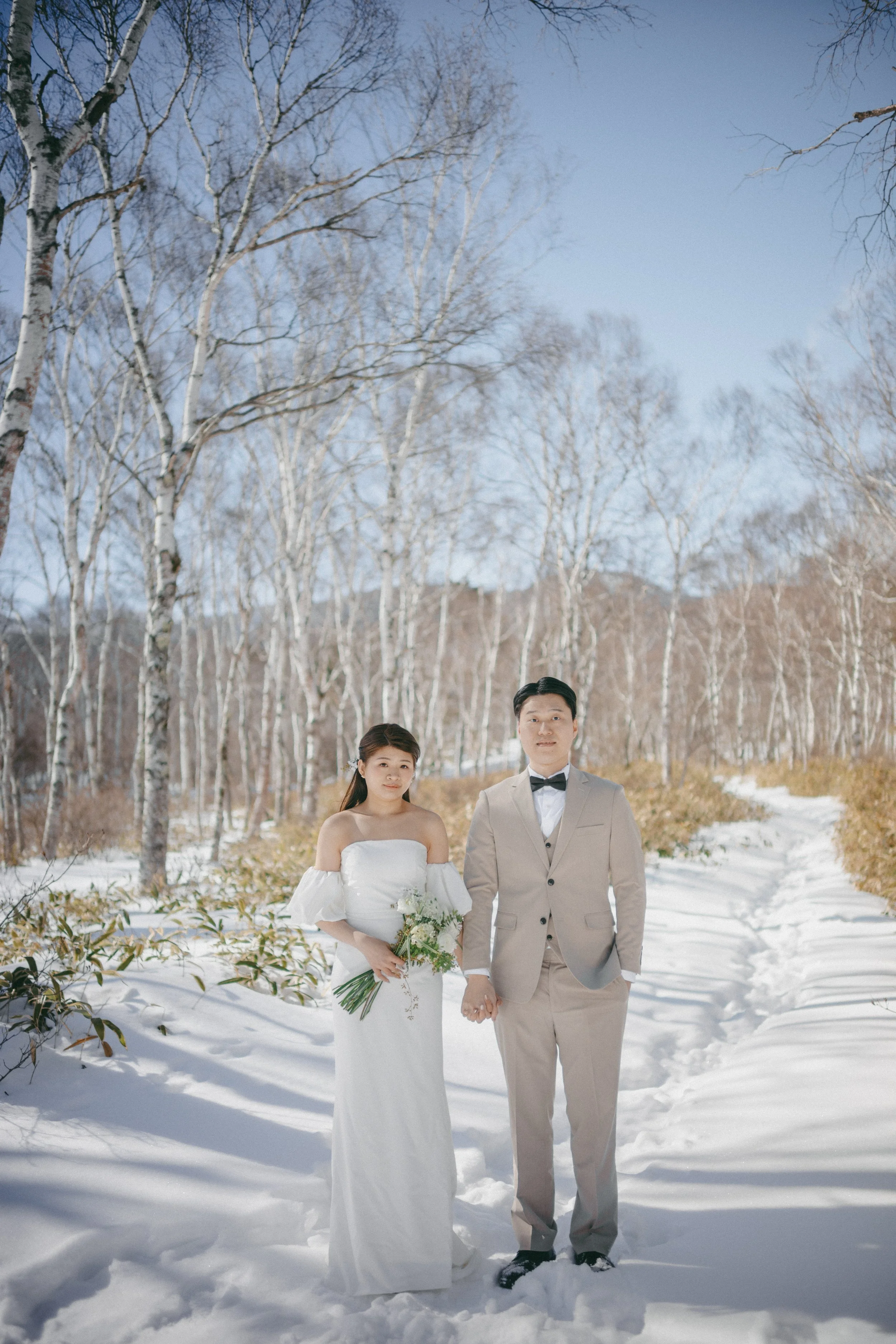 A bride in a white strapless wedding gown holding a bouquet stands next to a groom in a light gray suit and bow tie, holding hands, in a snowy outdoor setting with bare trees and a clear blue sky.