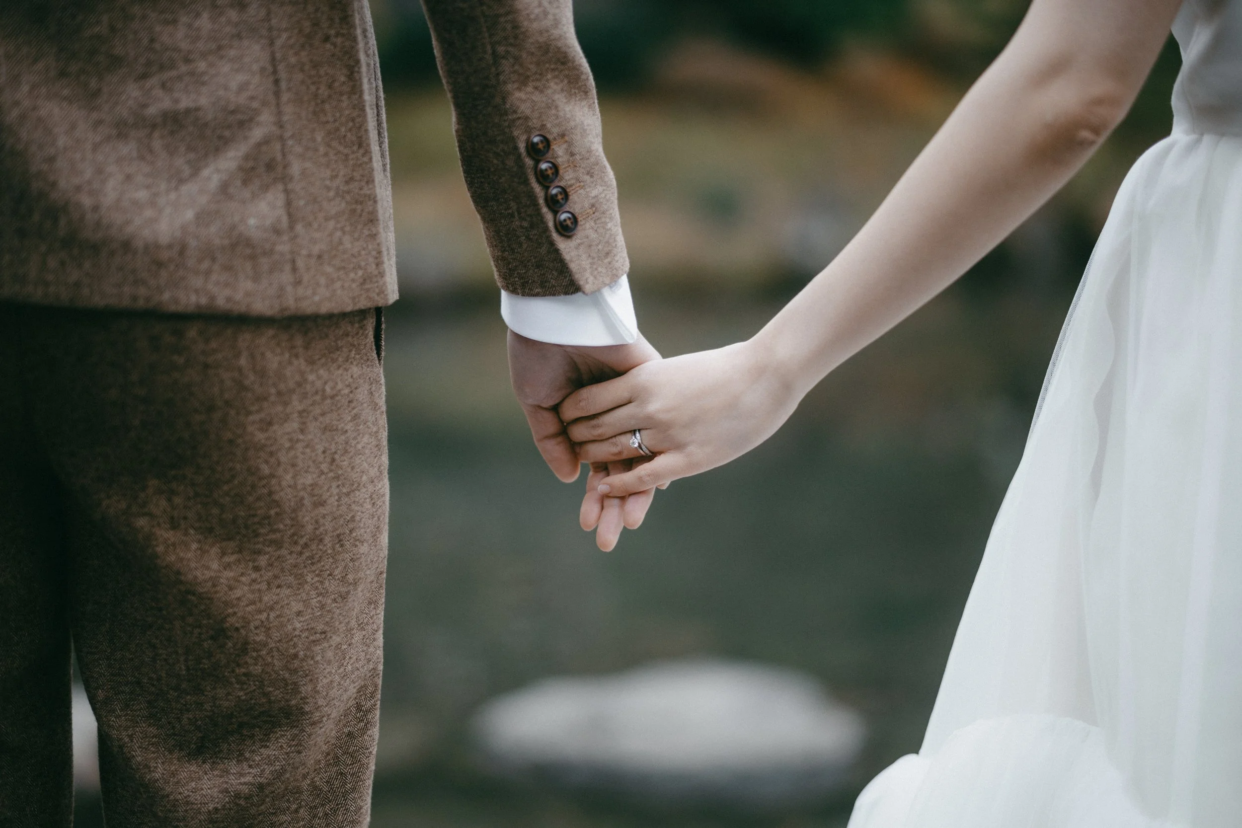 A close-up of a couple holding hands, with the woman displaying an engagement ring, outdoors with blurred background.