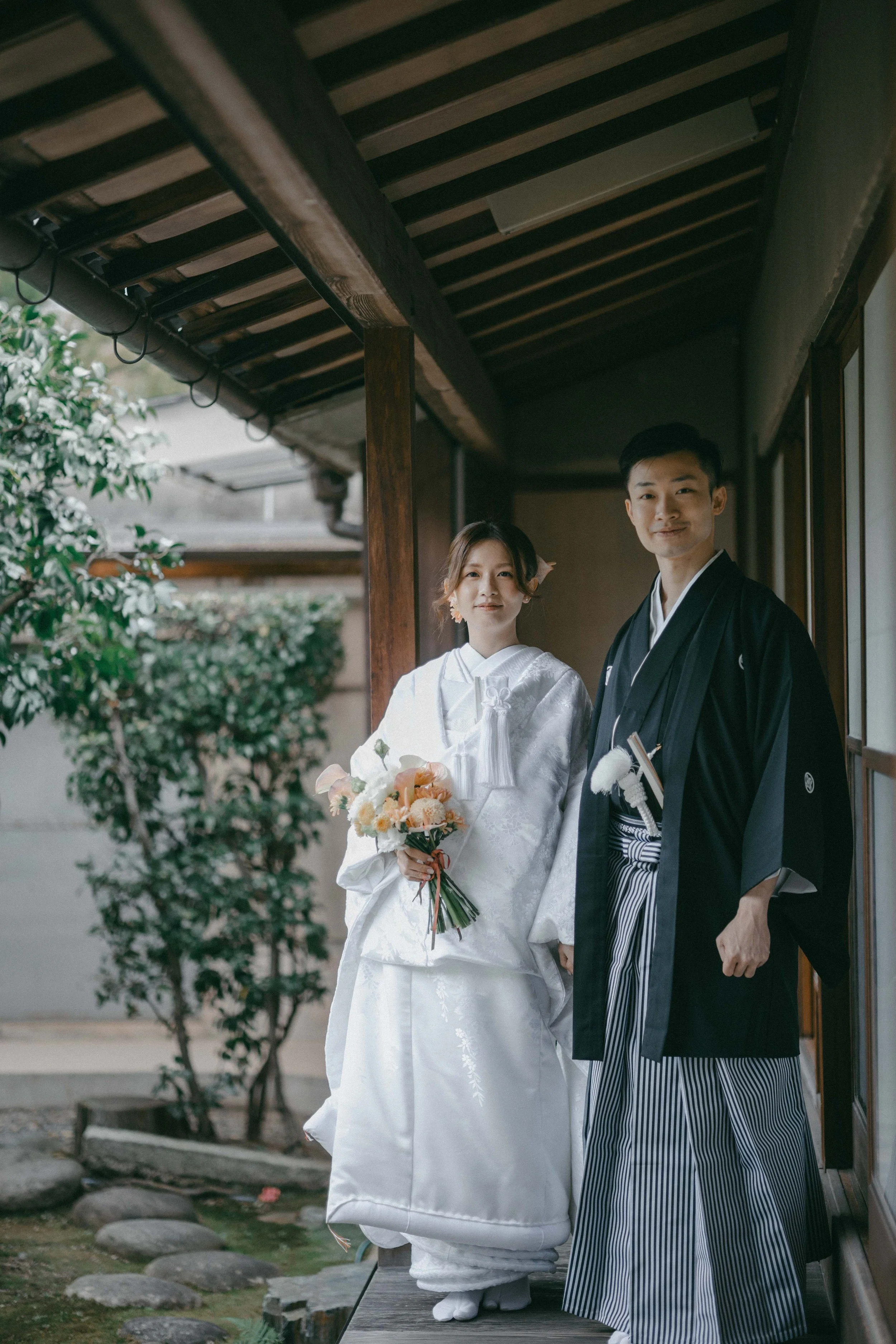 A bride and groom in traditional Japanese wedding attire standing on a wooden porch, holding hands, with the bride holding a bouquet of flowers.