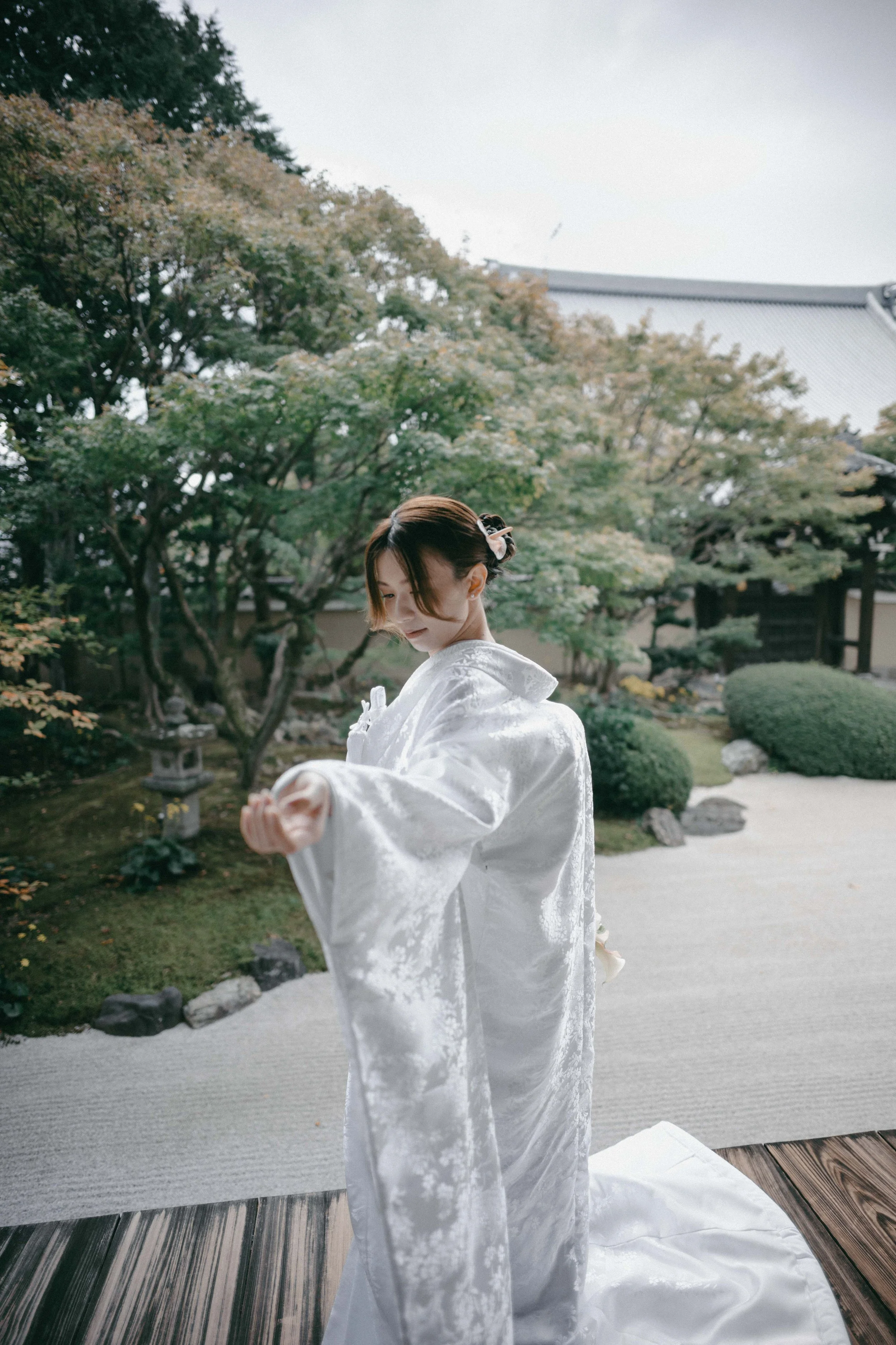 A woman in a white kimono standing on a wooden platform, with a traditional Japanese garden in the background, including trees, bushes, rocks, and a gravel path.