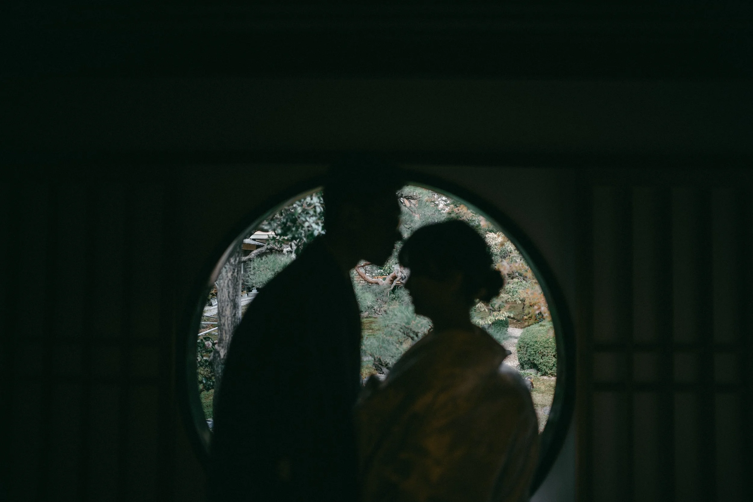 Silhouetted couple inside a traditional Japanese building looking at each other through a circular window, with a garden scene outside.