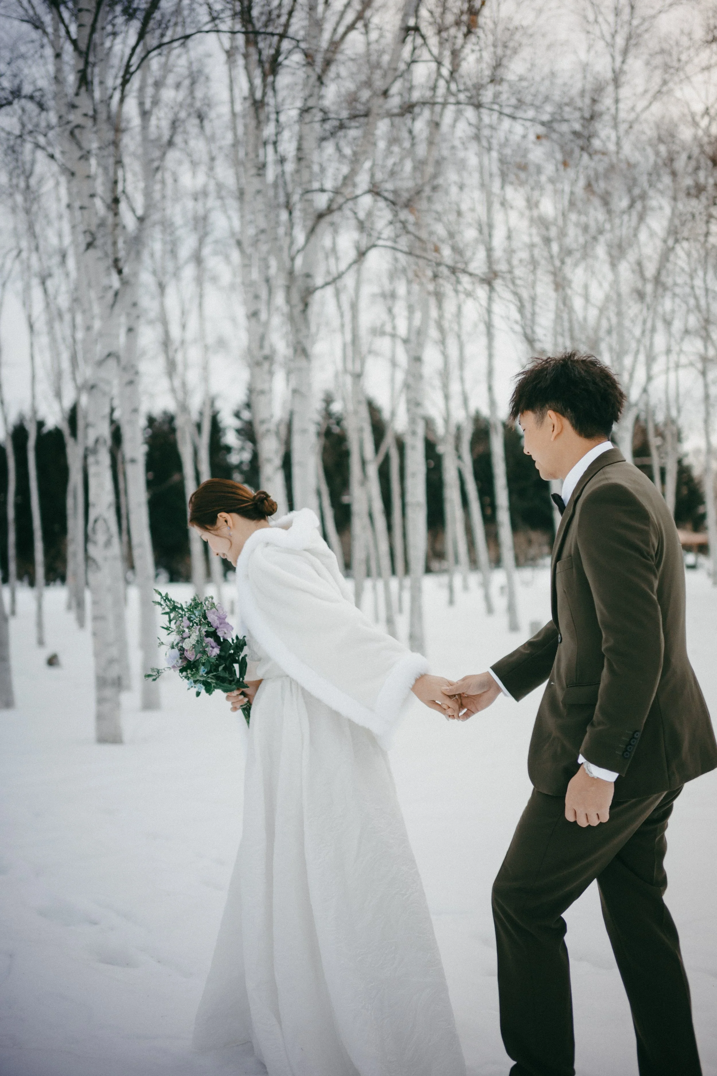Bride and groom holding hands in a snowy forest, bride holding a bouquet of flowers