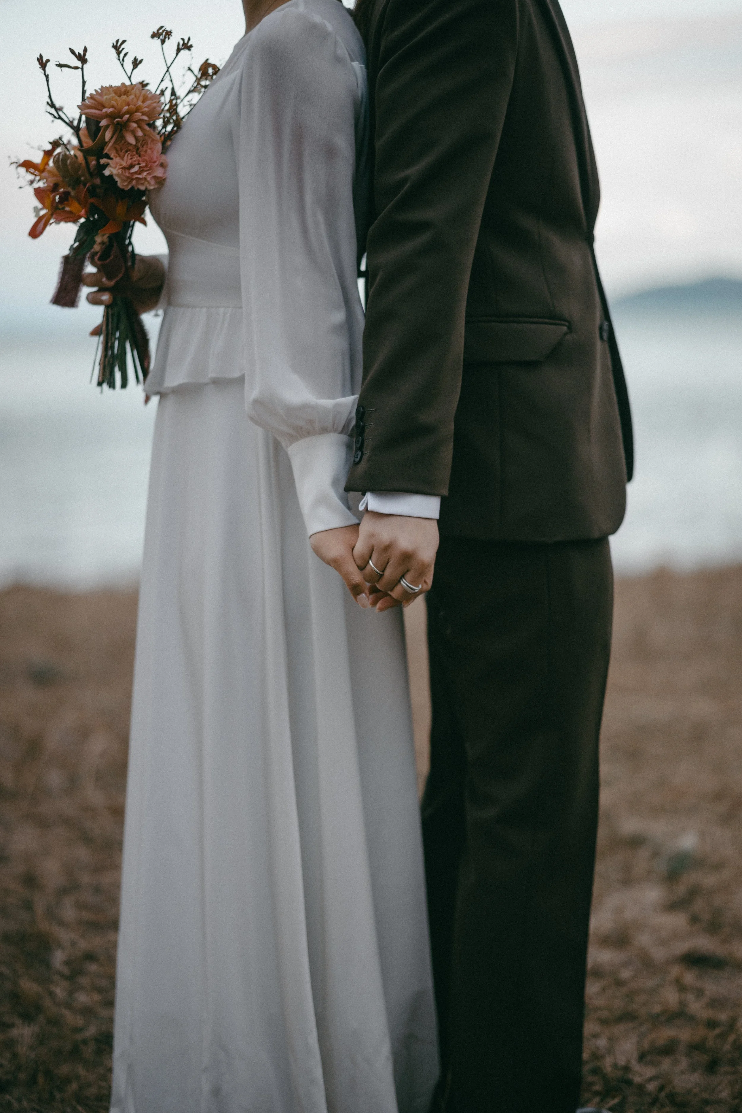 A bride and groom holding hands, standing outdoors near a body of water during sunset, with the bride holding a bouquet of flowers.
