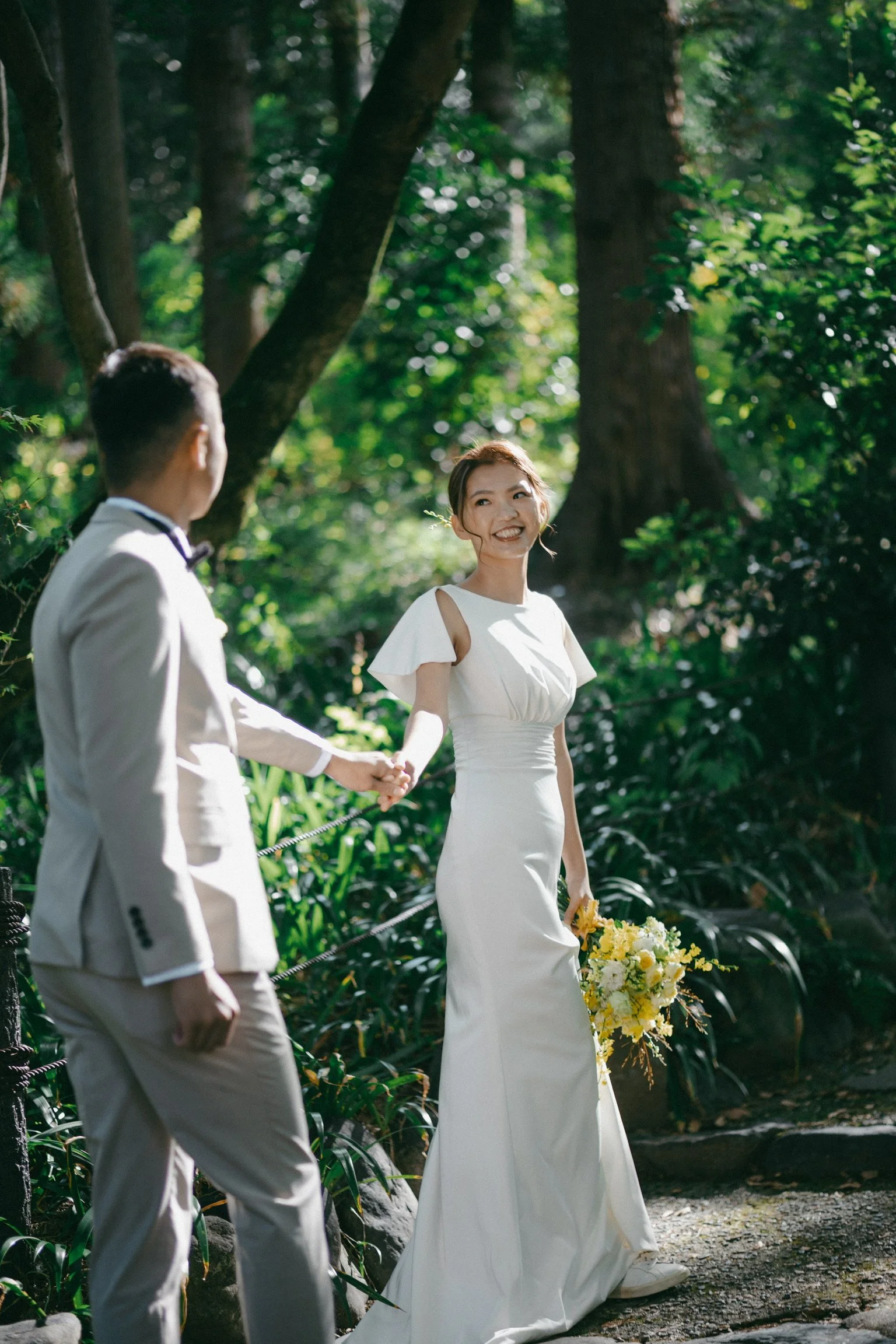A smiling bride in a white wedding gown holding a yellow bouquet, holding hands with a groom in a white suit, in a lush, green forest setting.