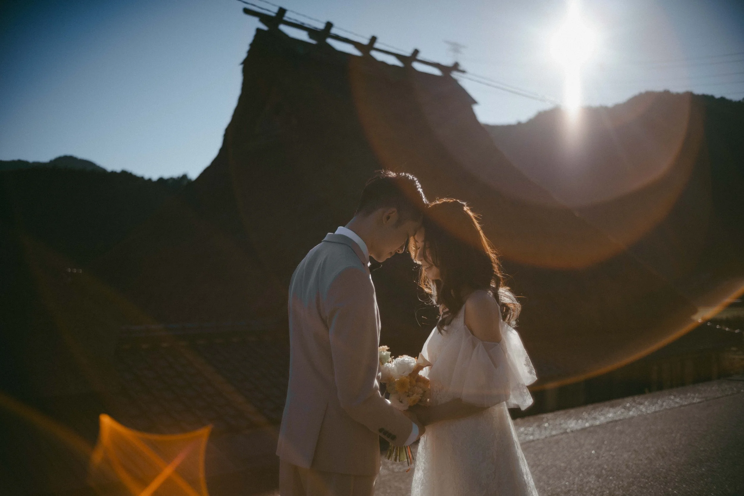 A bride and groom on their wedding day, standing close with foreheads touching, holding a bouquet of flowers outside during sunset with lens flare and mountain in the background.