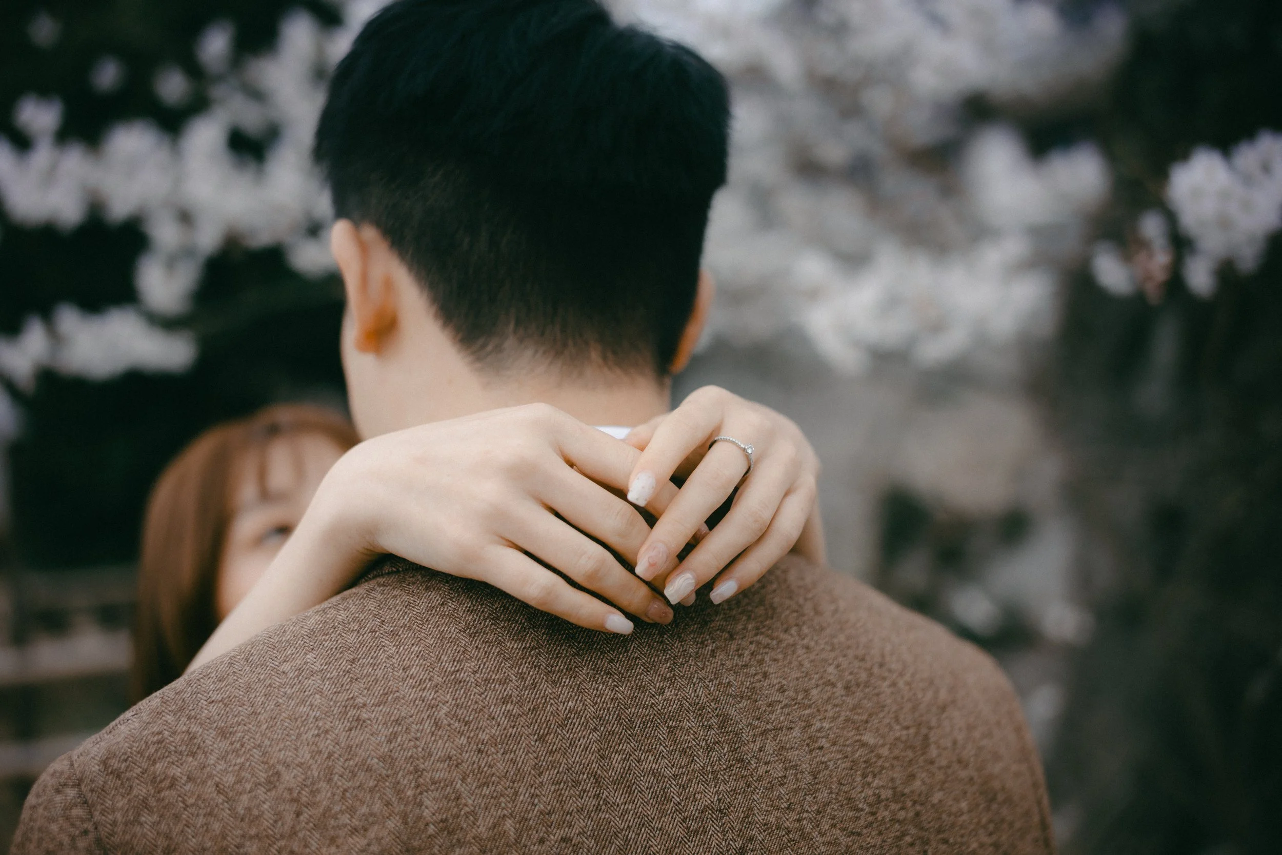 A woman embracing a man from behind, with her arms around his neck, and her hand resting on his shoulder, wearing a ring.