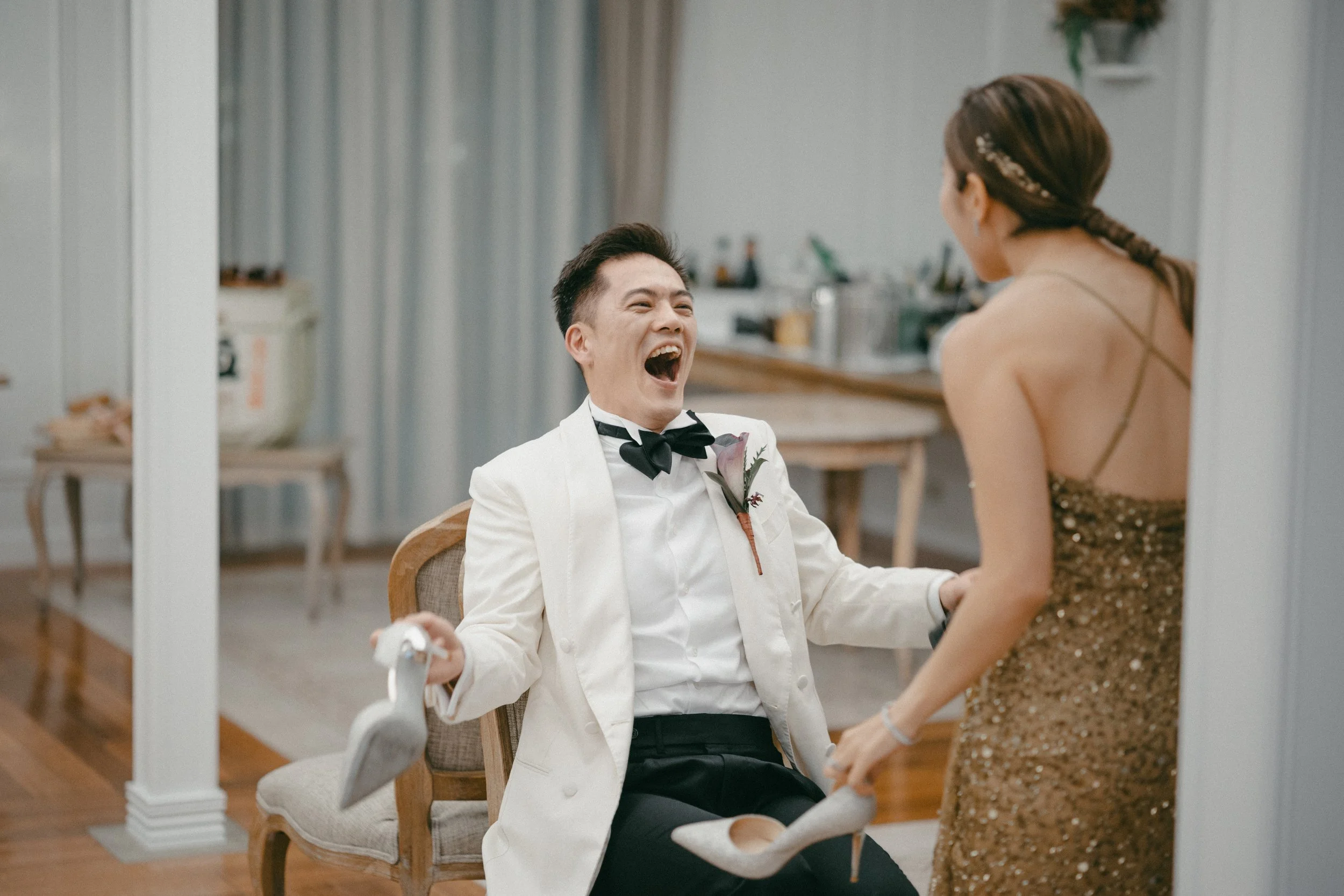 A man in a tuxedo laughing and holding a white high heel shoe while a woman in a gold dress stands in front of him, holding another high heel shoe, in a room with a wooden floor and a dining area in the background.