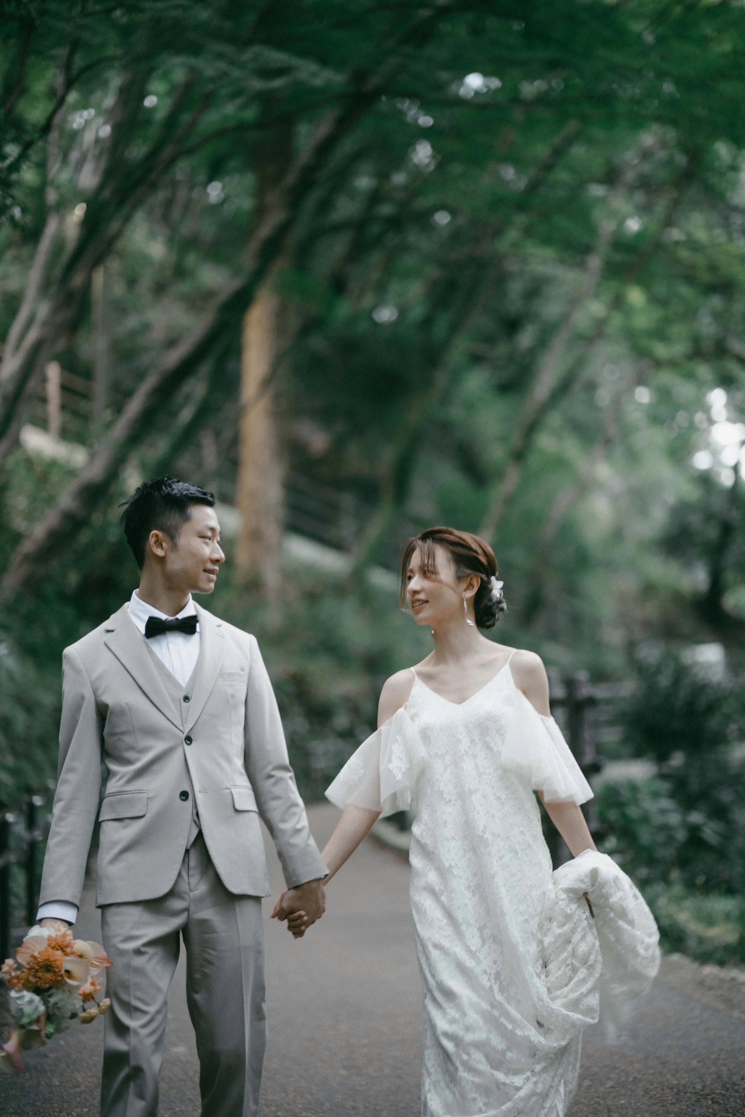 A bride and groom walking hand in hand outdoors in a wooded area, smiling at each other.