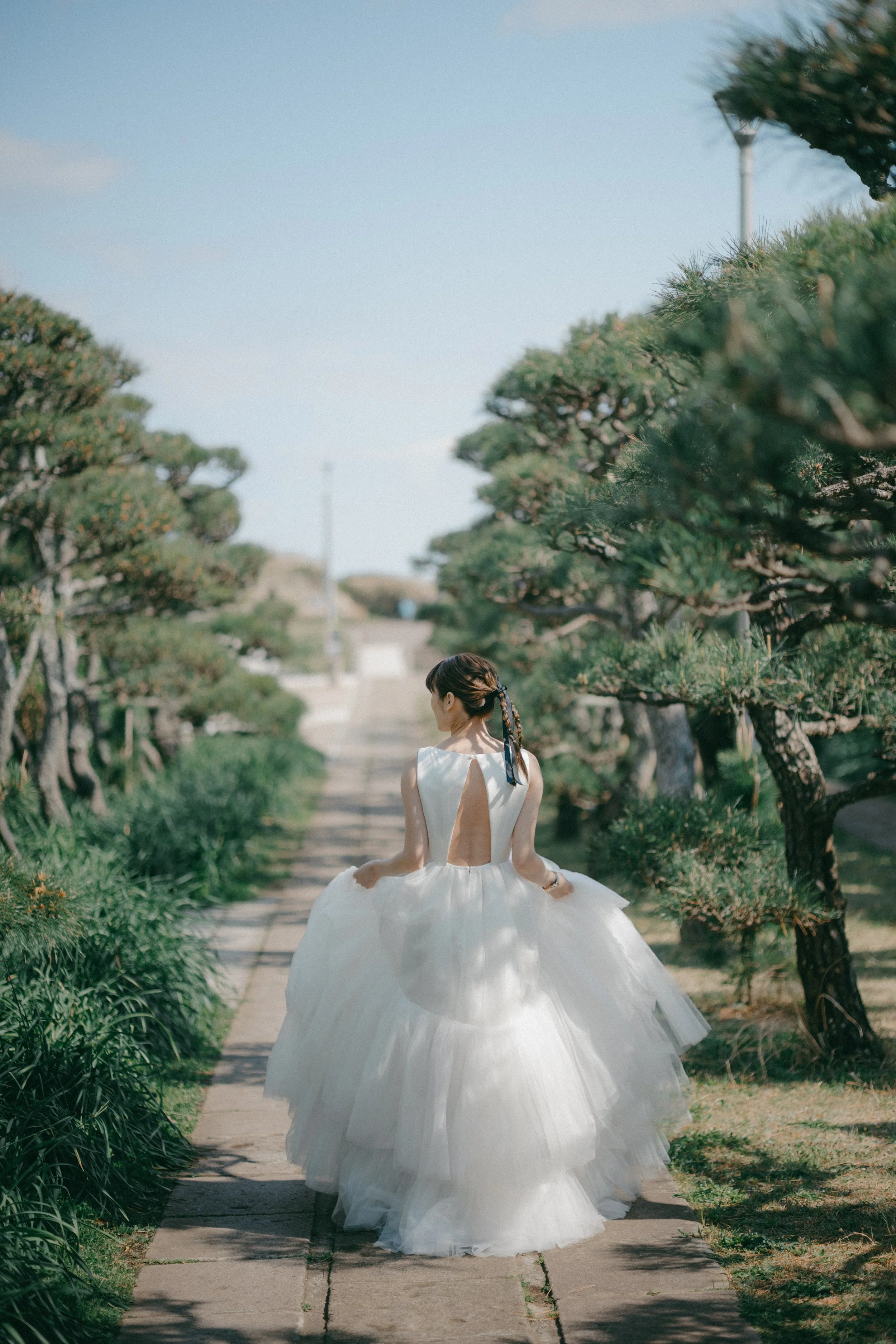 A woman in a white dress with an open back walking along a stone path surrounded by green trees.