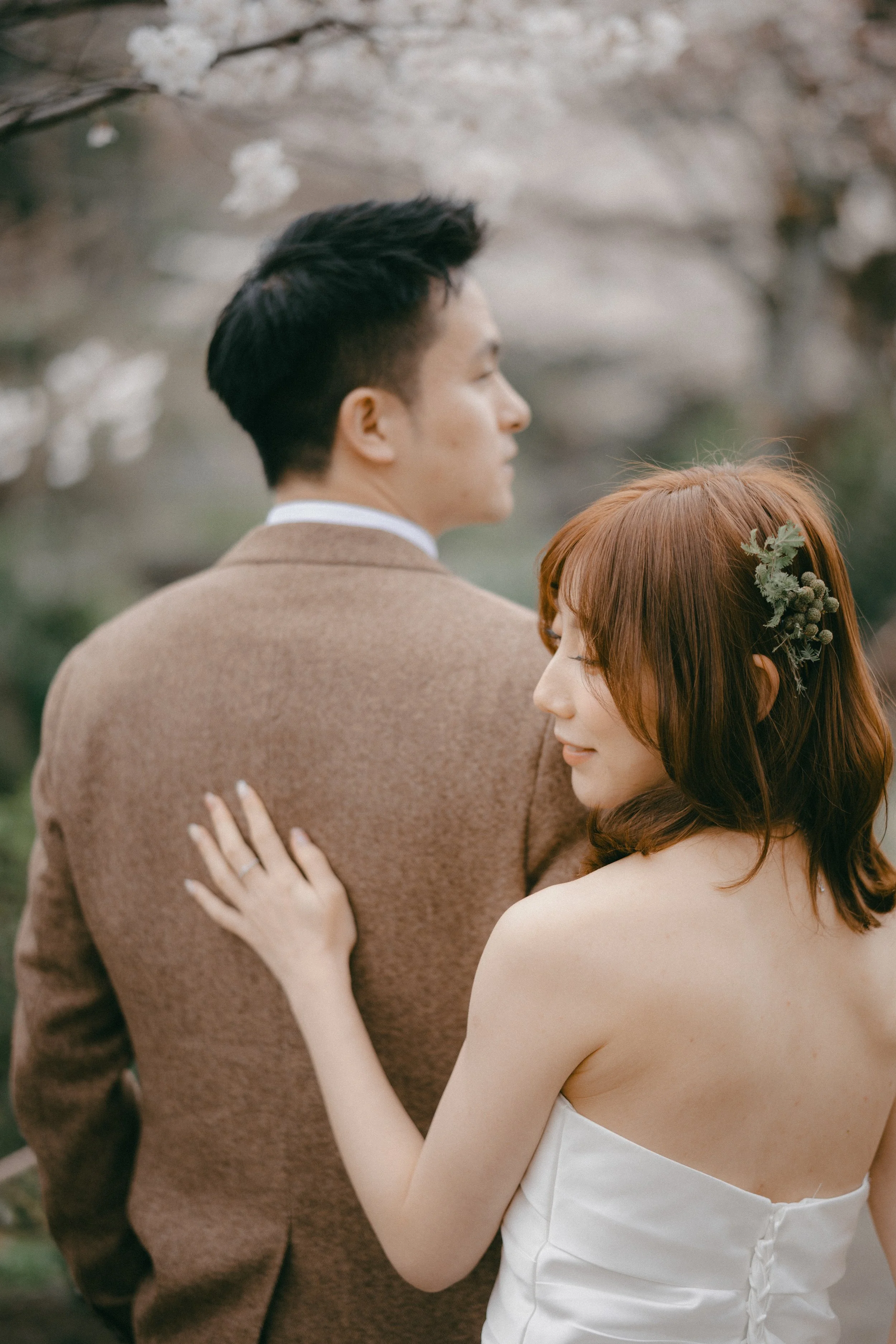 A woman with red hair and a flower hair accessory embraces a man in a brown suit outdoors.