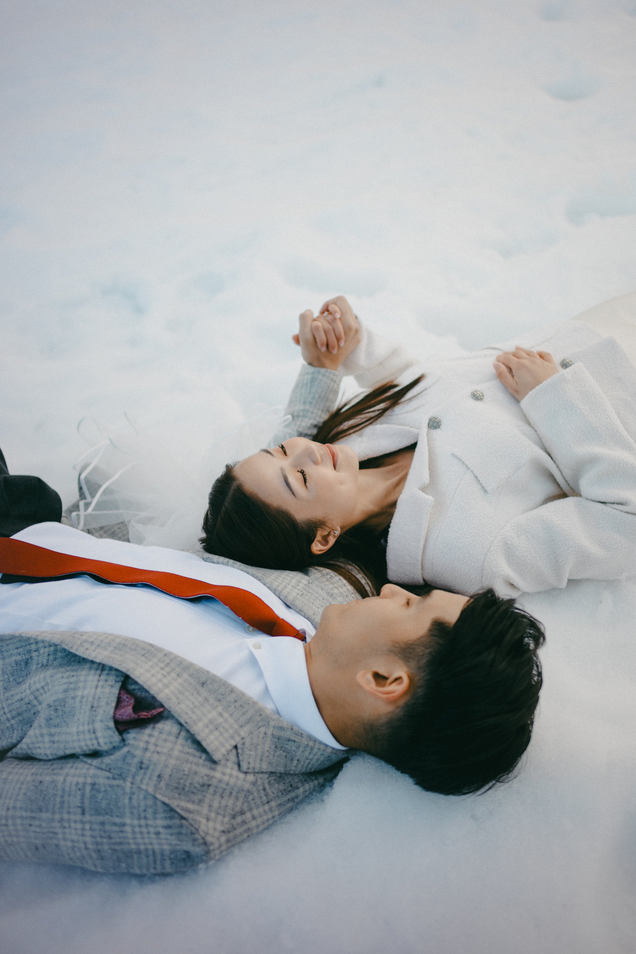 A young couple lying on the snow, holding hands and gazing at each other in a winter setting.