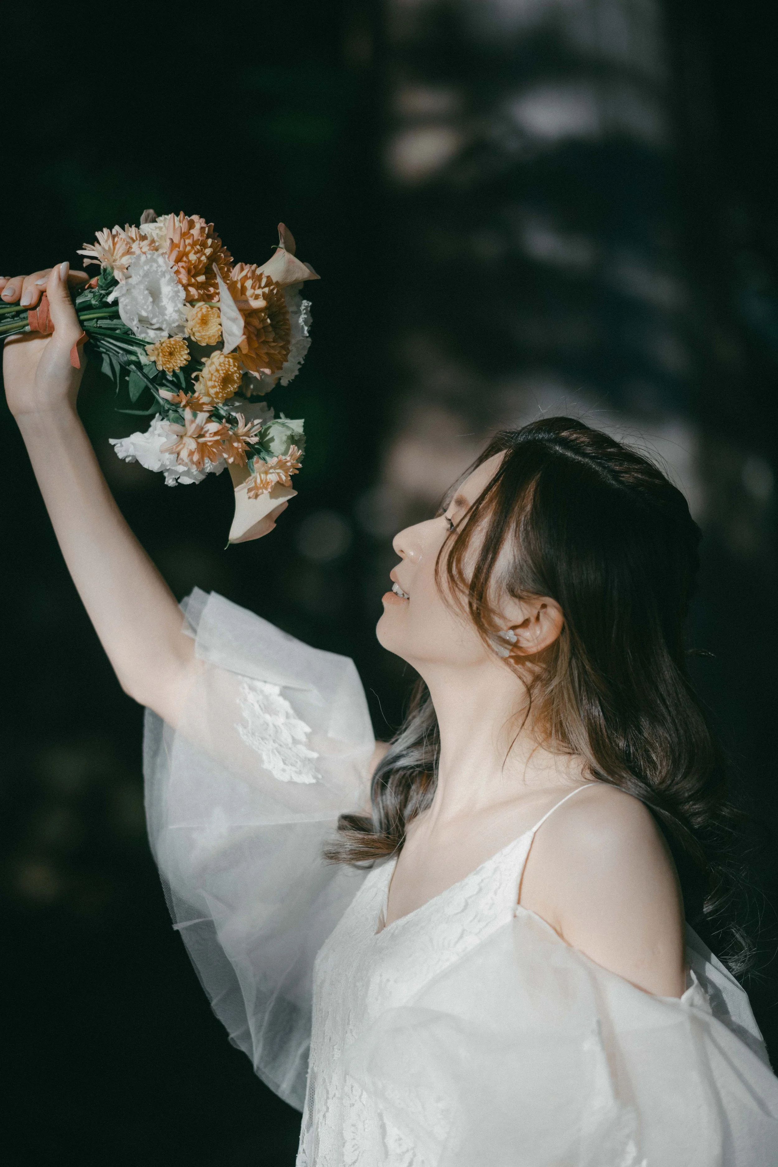 A woman in a white dress holds a bouquet of flowers and looks at it smiling outdoors.