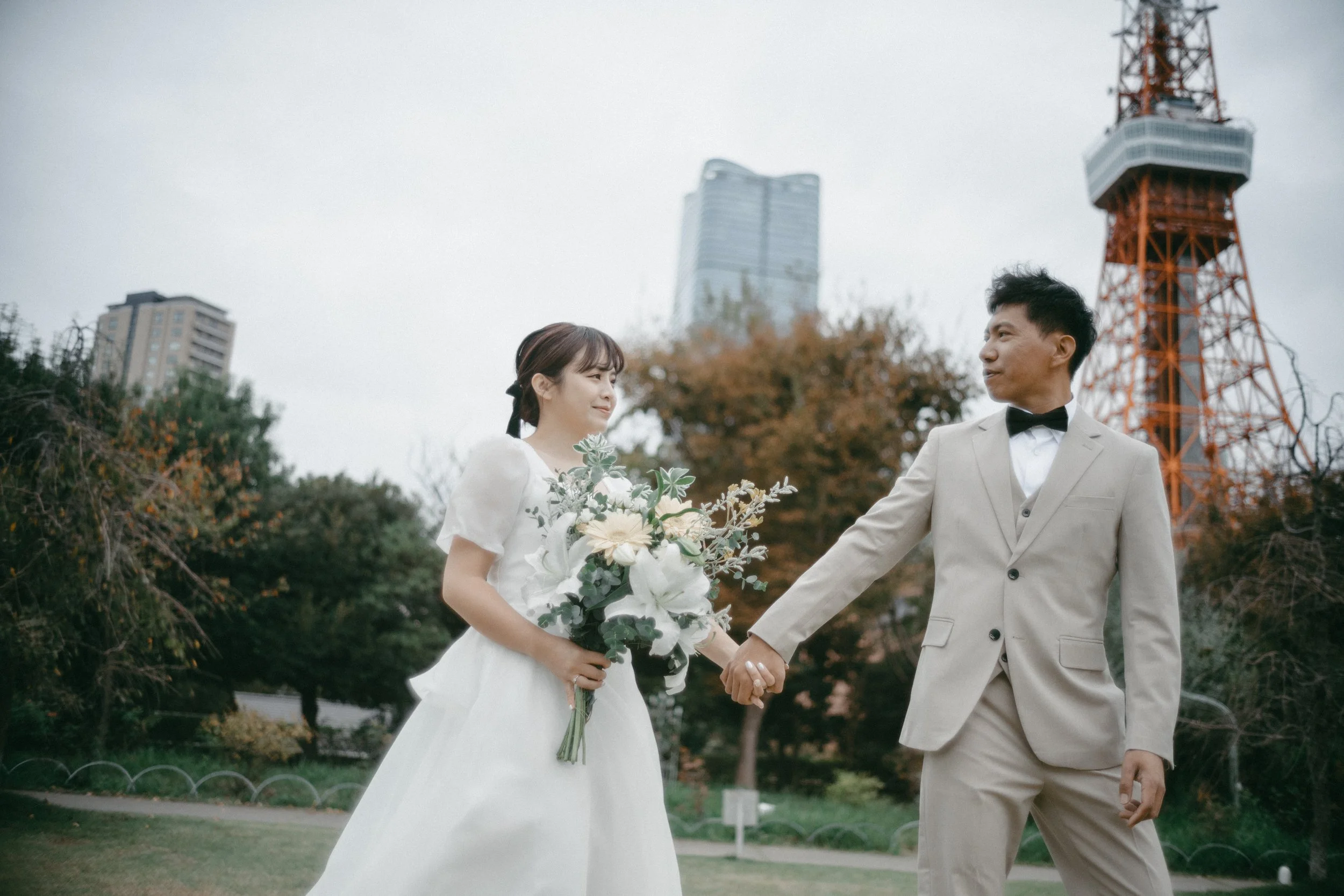 A bride and groom holding hands outdoors in a park, with the bride holding a bouquet of white flowers and greenery, and a cityscape with a tall communication tower in the background.