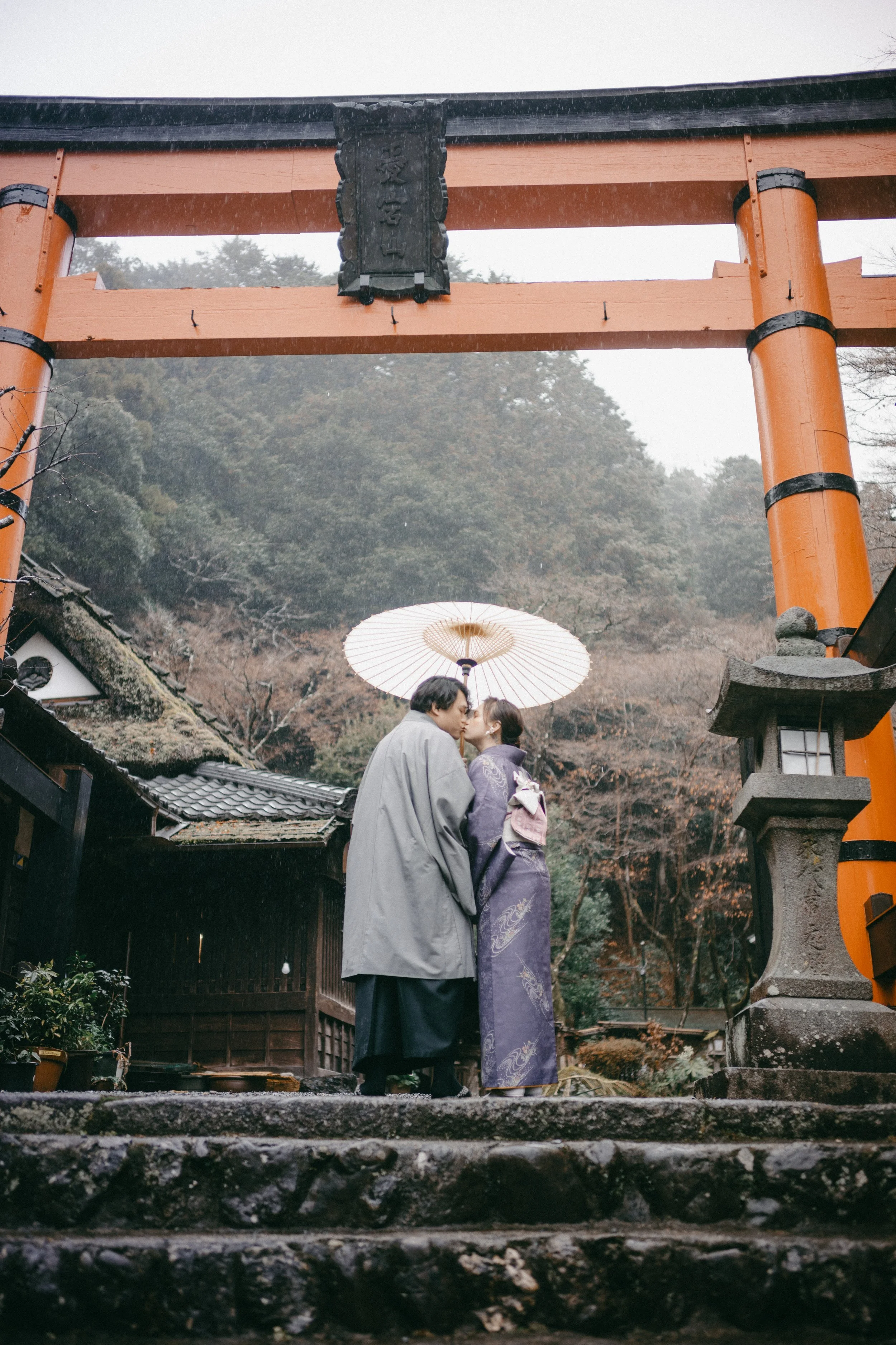 A couple dressed in traditional Japanese clothing sharing a kiss under a paper parasol, standing on stone steps in front of a torii gate at a Japanese shrine.