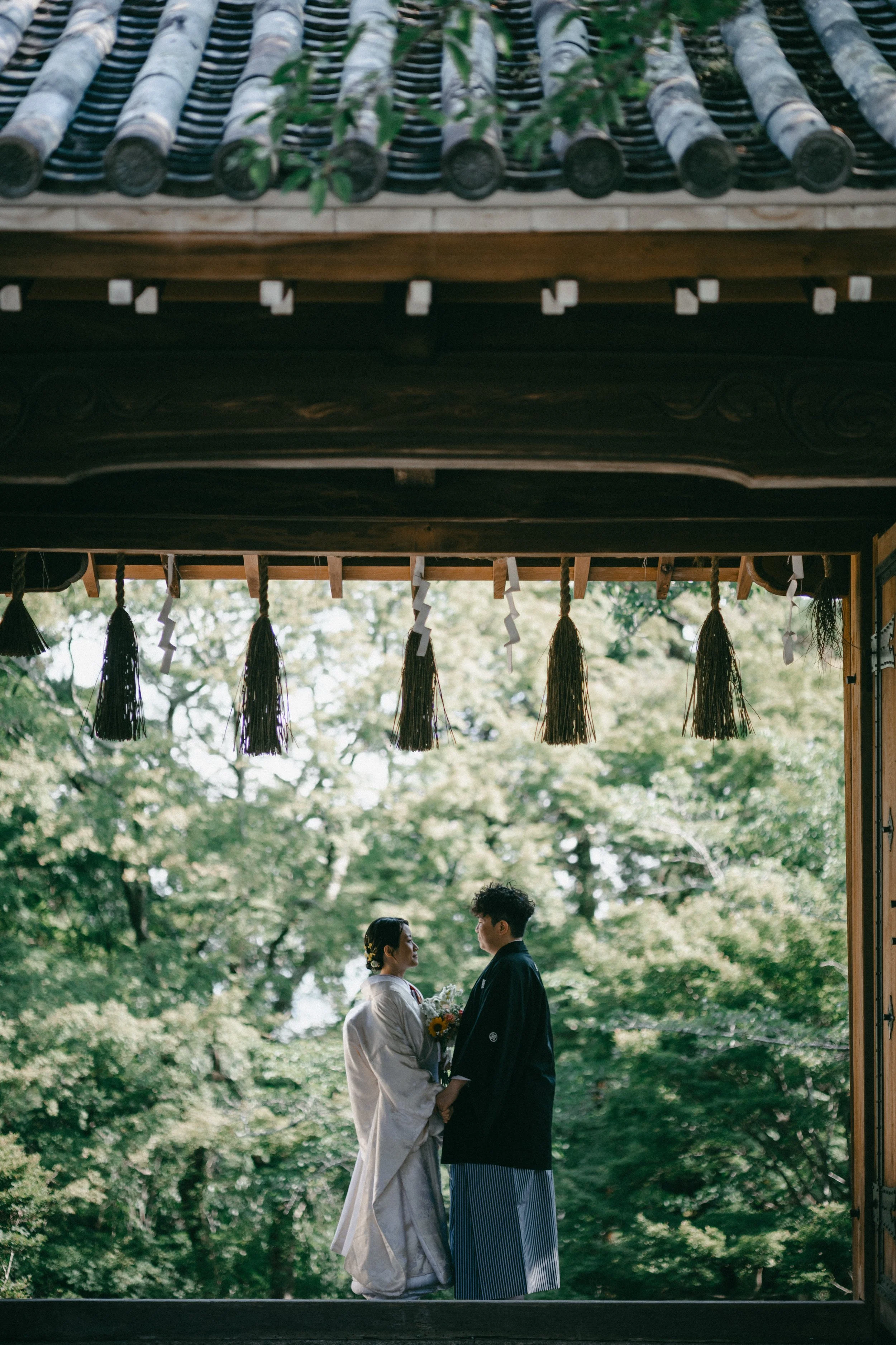 A bride and groom in traditional wedding attire holding hands and facing each other under a wooden shrine with a nature background.