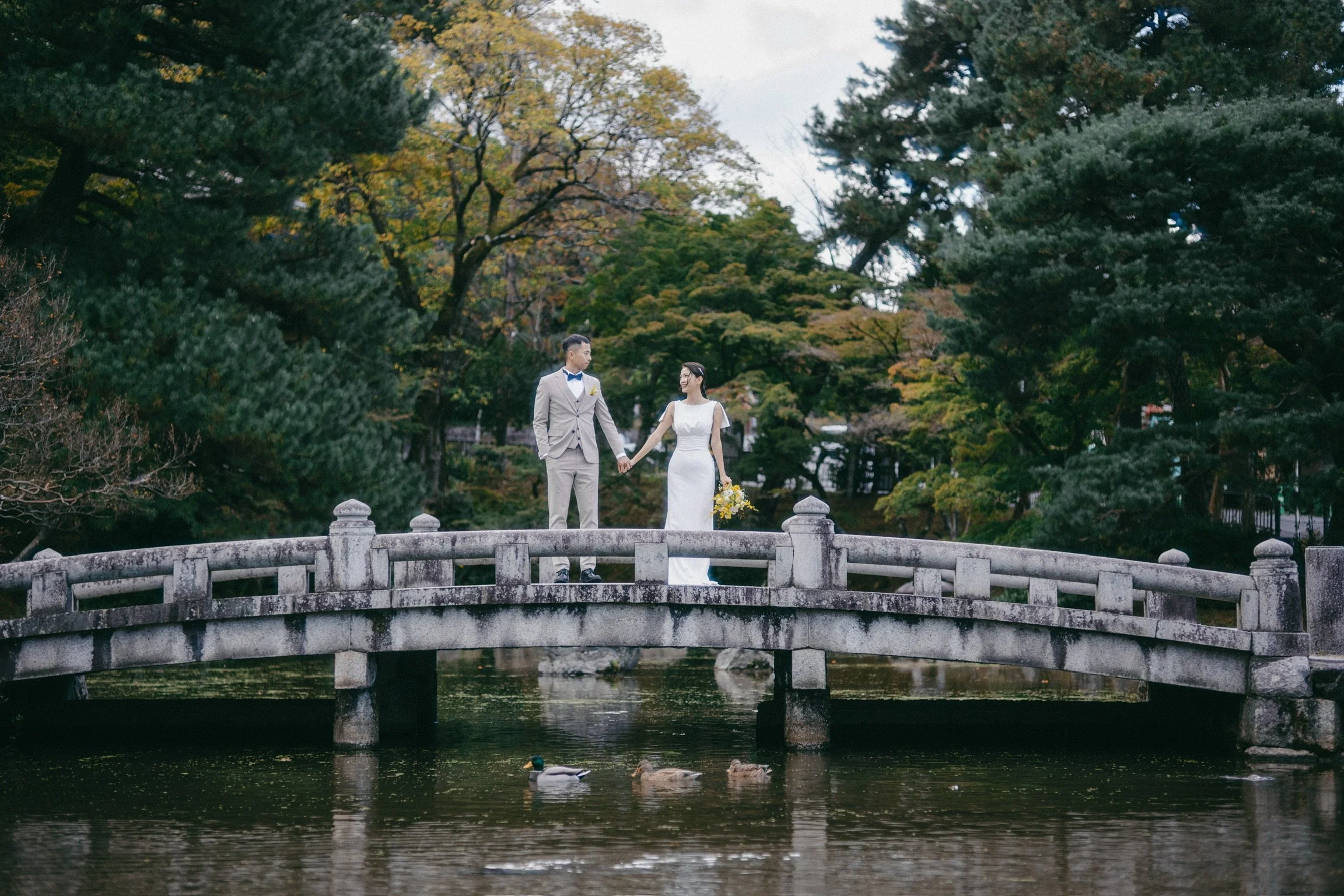 A bride and groom holding hands on a stone bridge over a pond in a park, surrounded by trees with fall foliage, with ducks swimming below.