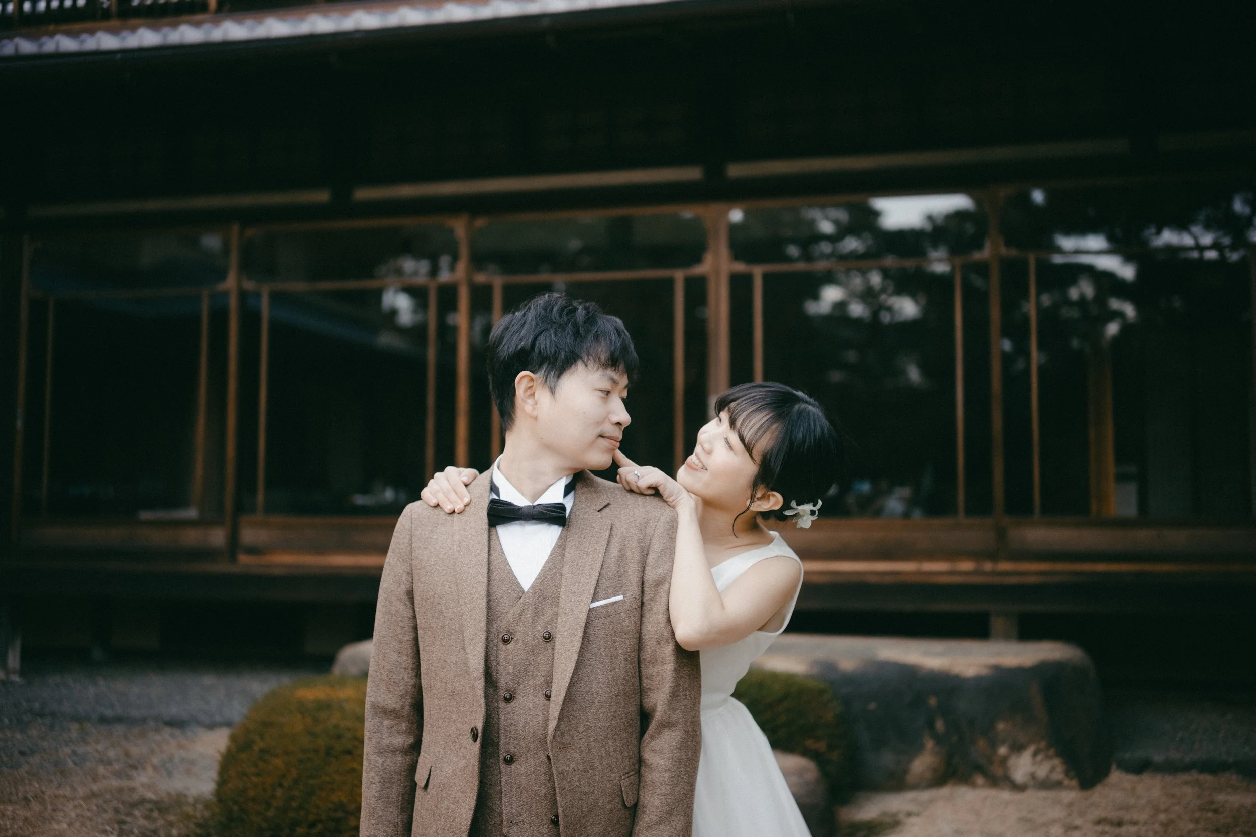 A young couple dressed in wedding attire standing close together outdoors, with the woman touching the man's shoulder and smiling at him. The man is wearing a brown suit with a black bow tie, and the woman is wearing a white wedding dress with a flow
