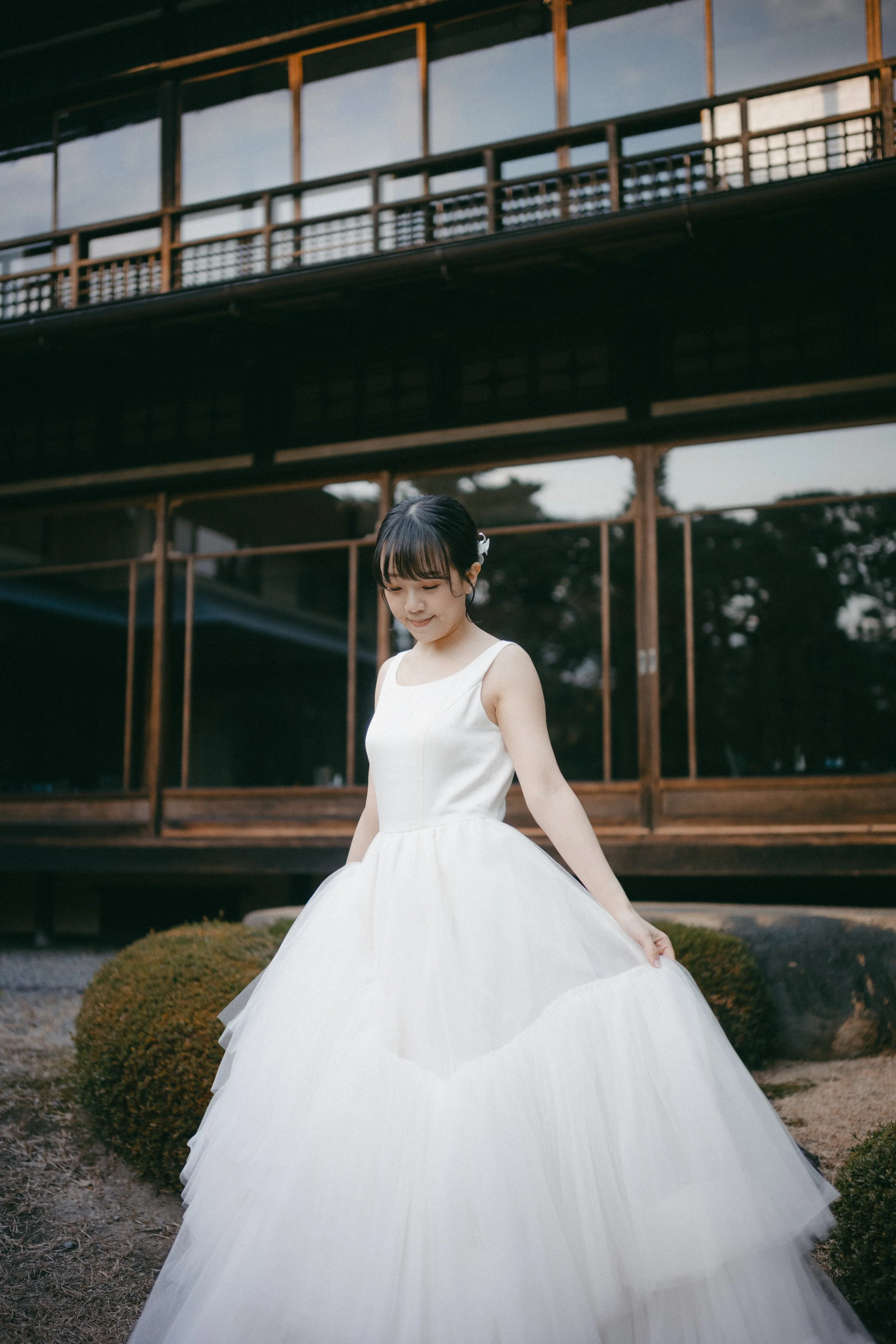 A young woman in a white wedding dress standing outdoors in front of a traditional wooden building, with shrubs and rocks visible around her.