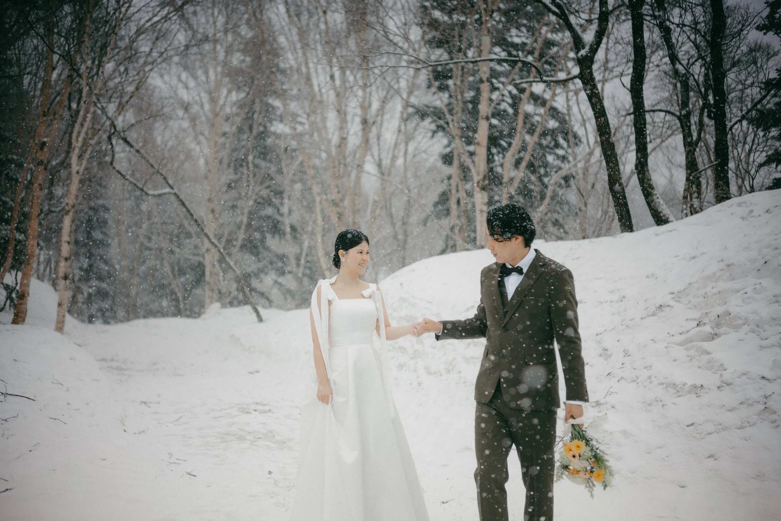 A bride and groom holding hands in a snowy forest, with snow falling around them. The bride is in a white gown, and the groom is in a dark suit with a bow tie, holding a bouquet of flowers.