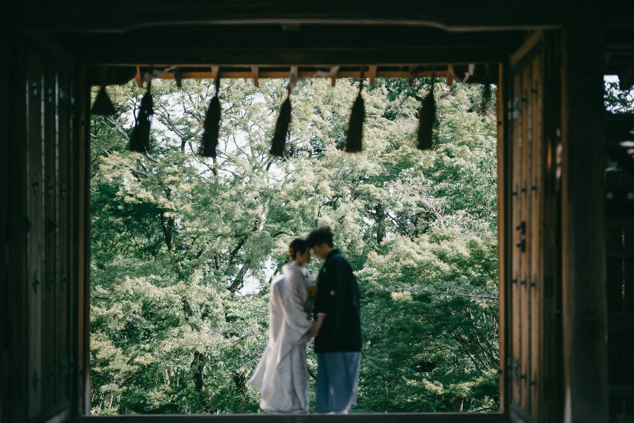 A couple dressed in traditional Japanese wedding attire standing close together outdoors, framed by a wooden structure with lush green trees in the background.