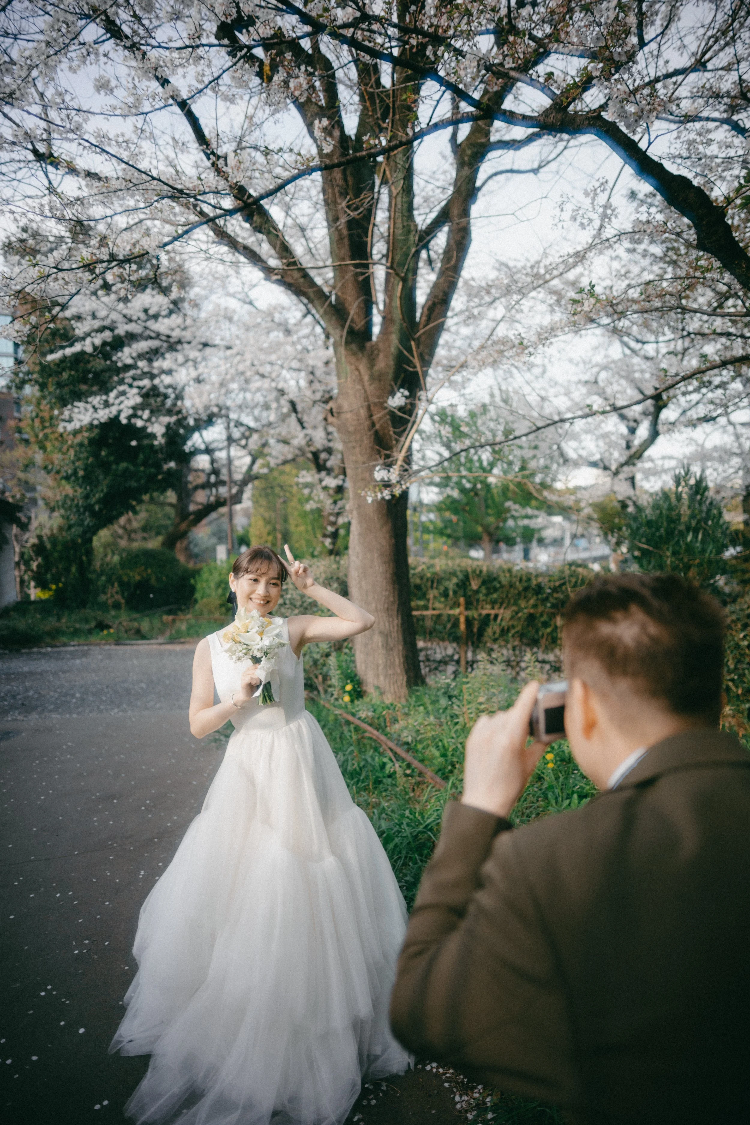A bride in a white wedding dress takes a photo with a man in a suit under blooming cherry blossom trees during daytime.