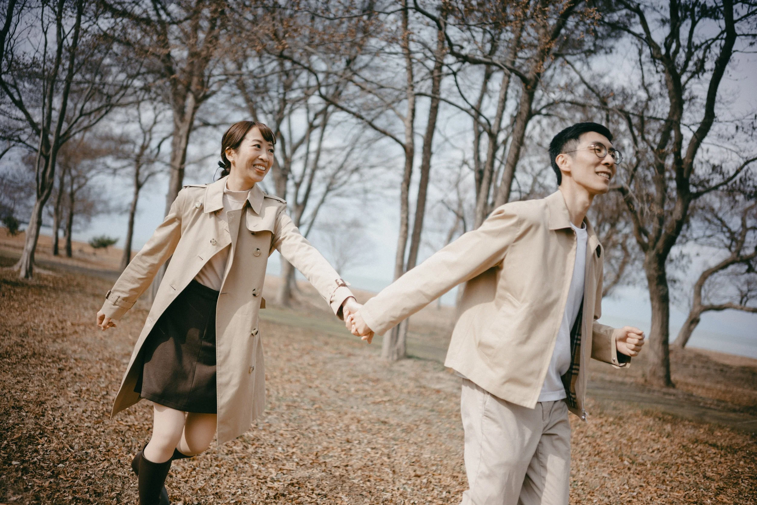 A smiling couple holding hands and running through a park with bare trees during autumn.
