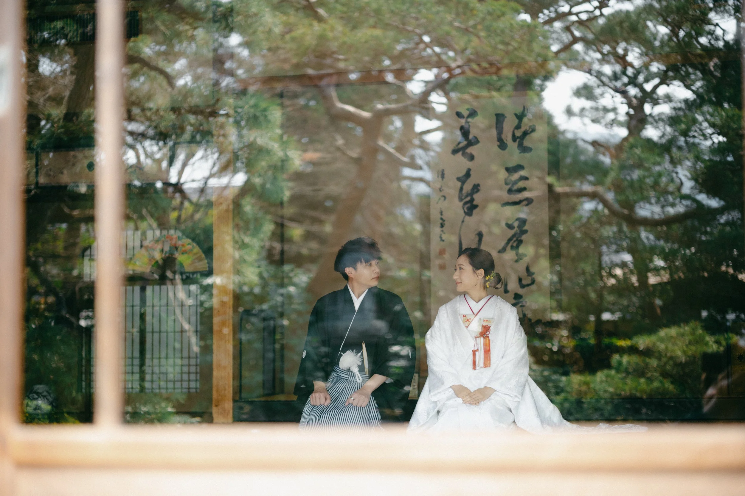 A couple in traditional Japanese wedding attire sitting and looking at each other through a window, with trees and Japanese calligraphy in the background.