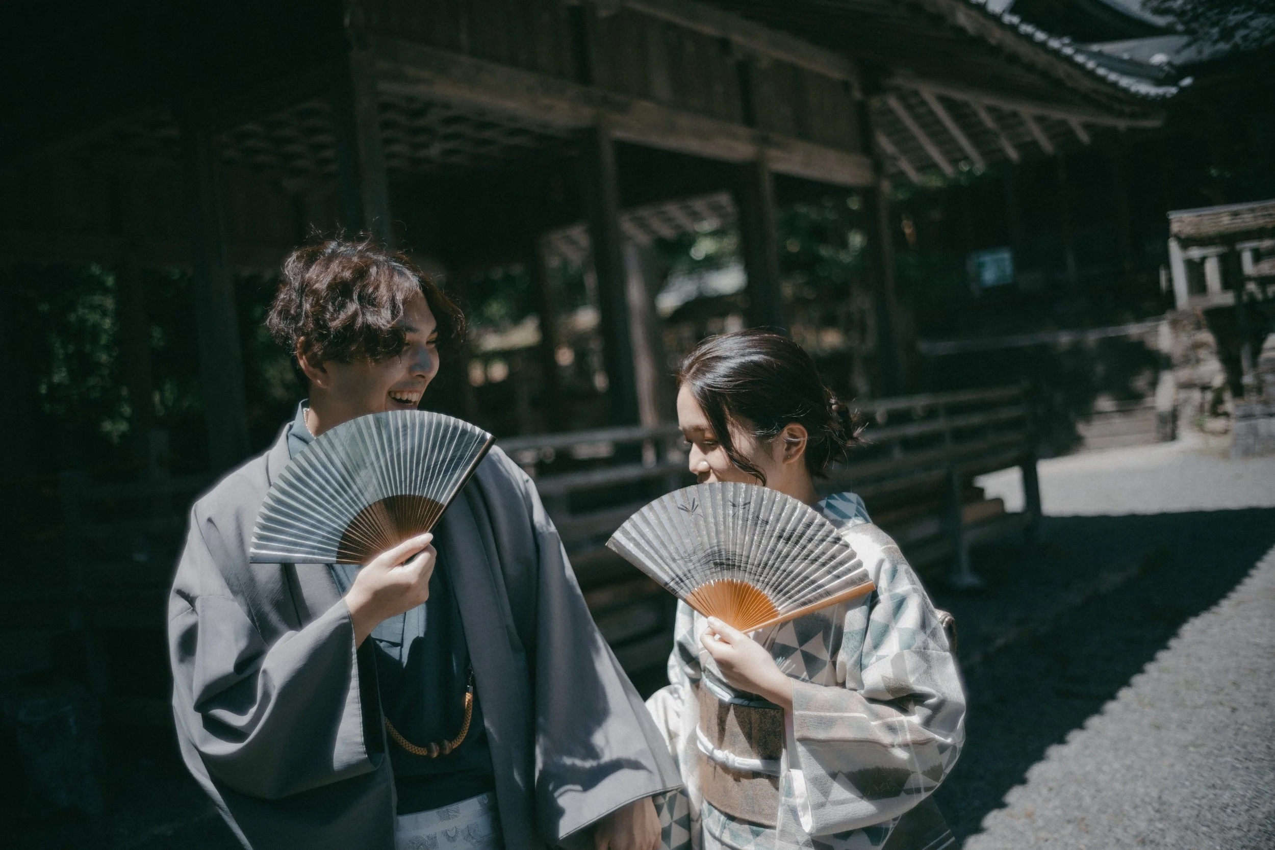 Two women wearing traditional Japanese kimonos holding folding fans and smiling at each other outdoors.