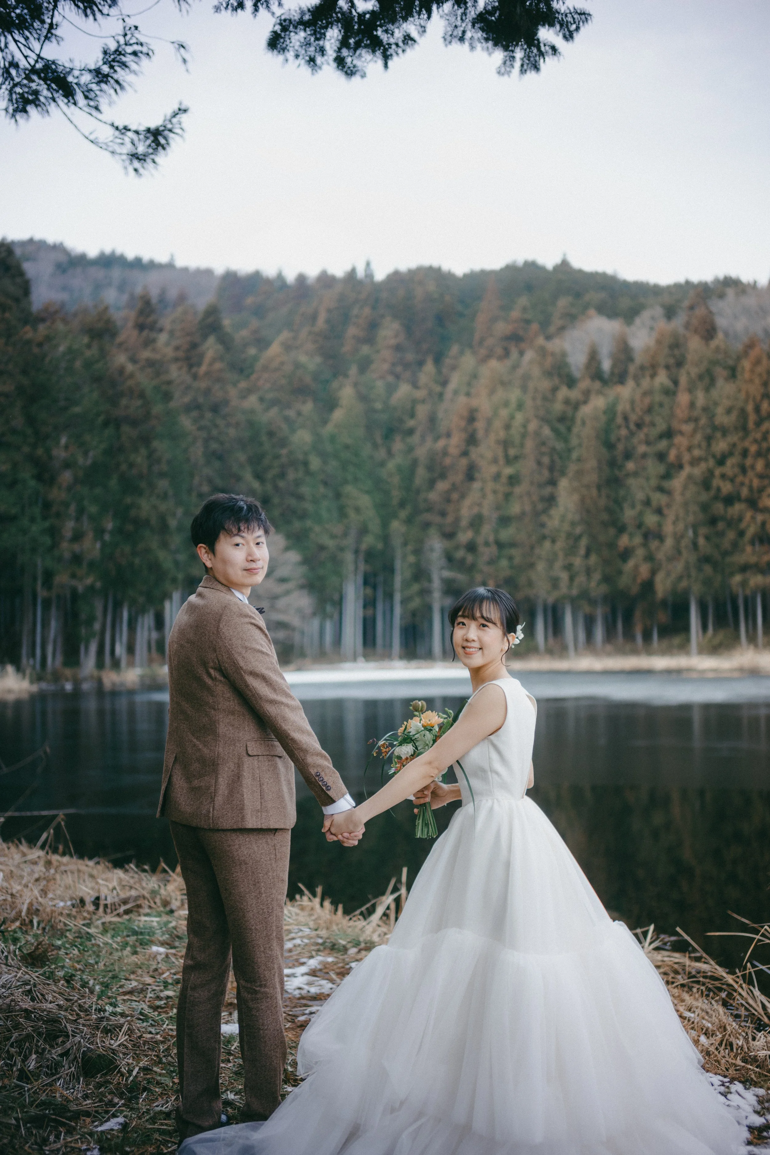 A bride and groom standing hand-in-hand outdoors by a lake surrounded by trees, with mountains in the background.