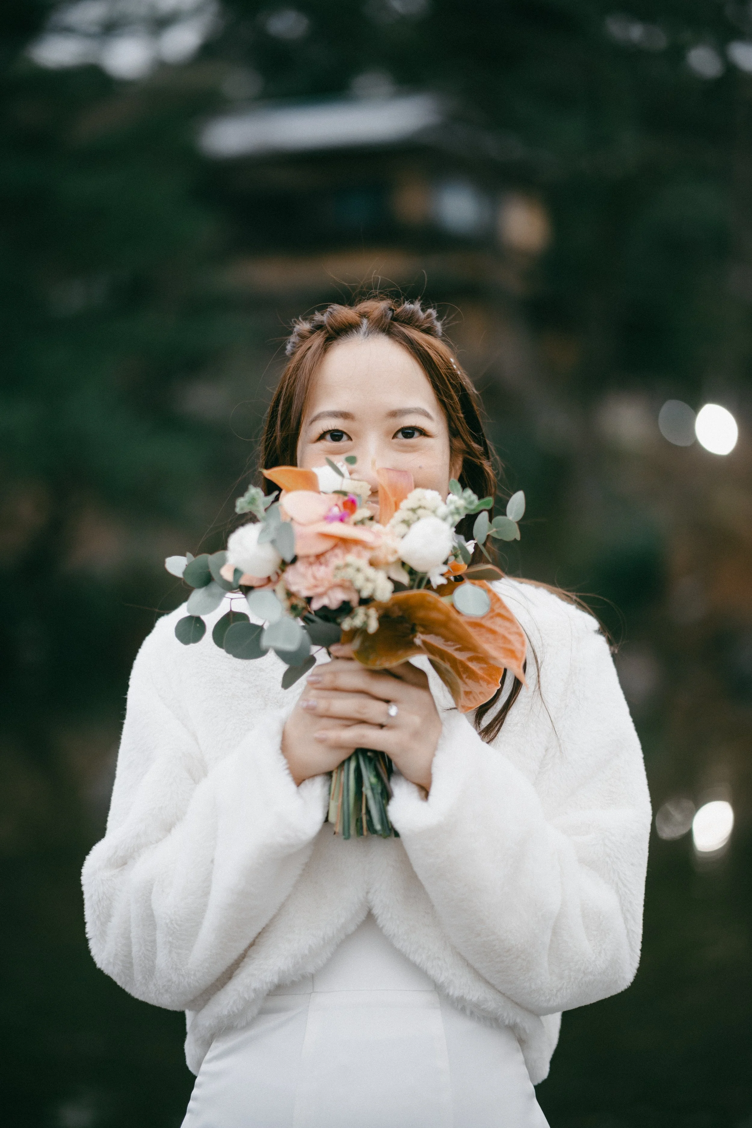 A woman in a white coat holding a bouquet of flowers in front of her face, with only her eyes visible, standing outdoors with dark green trees in the background.