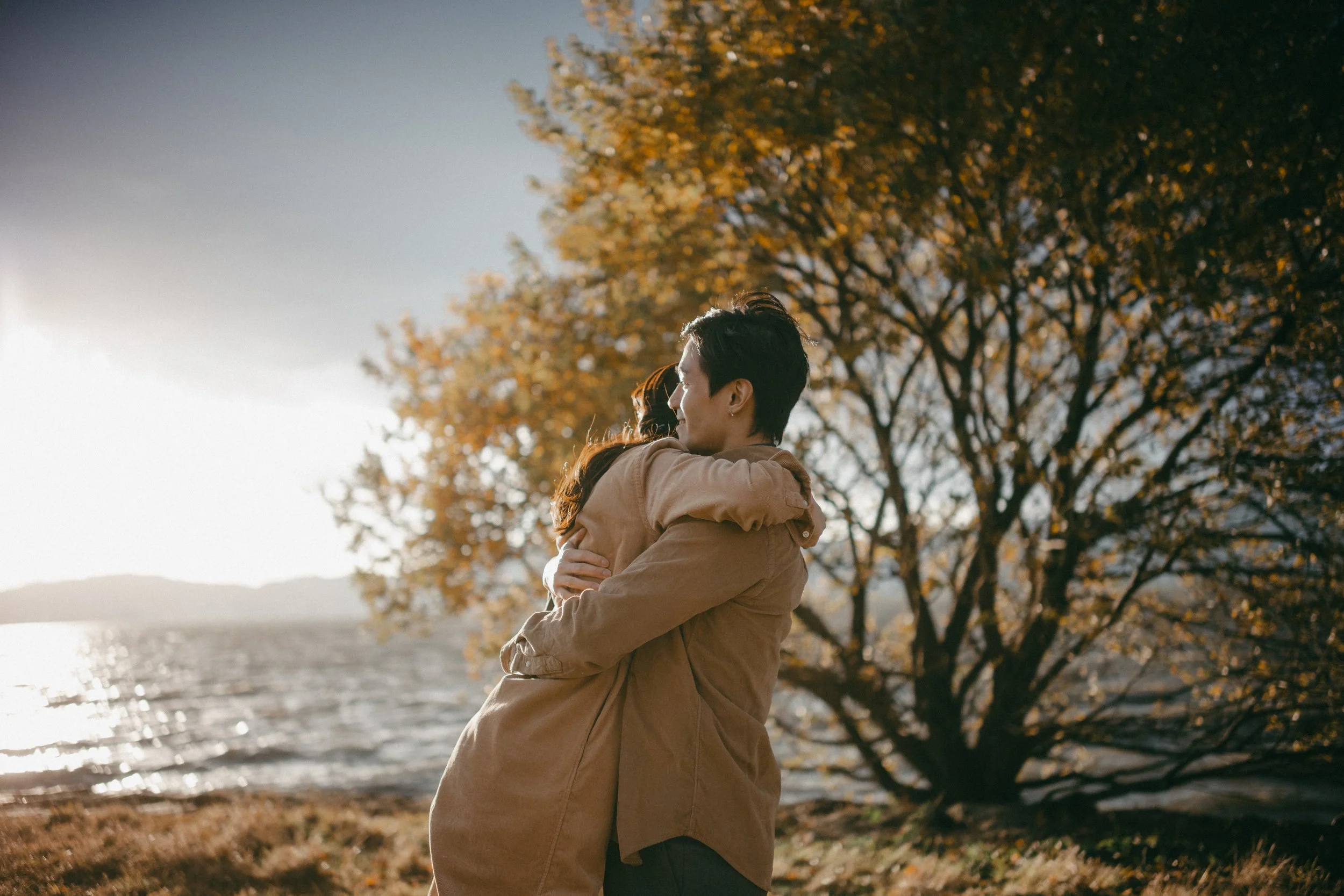 A couple hugging by a lake during autumn, with a large tree with orange leaves in the background.